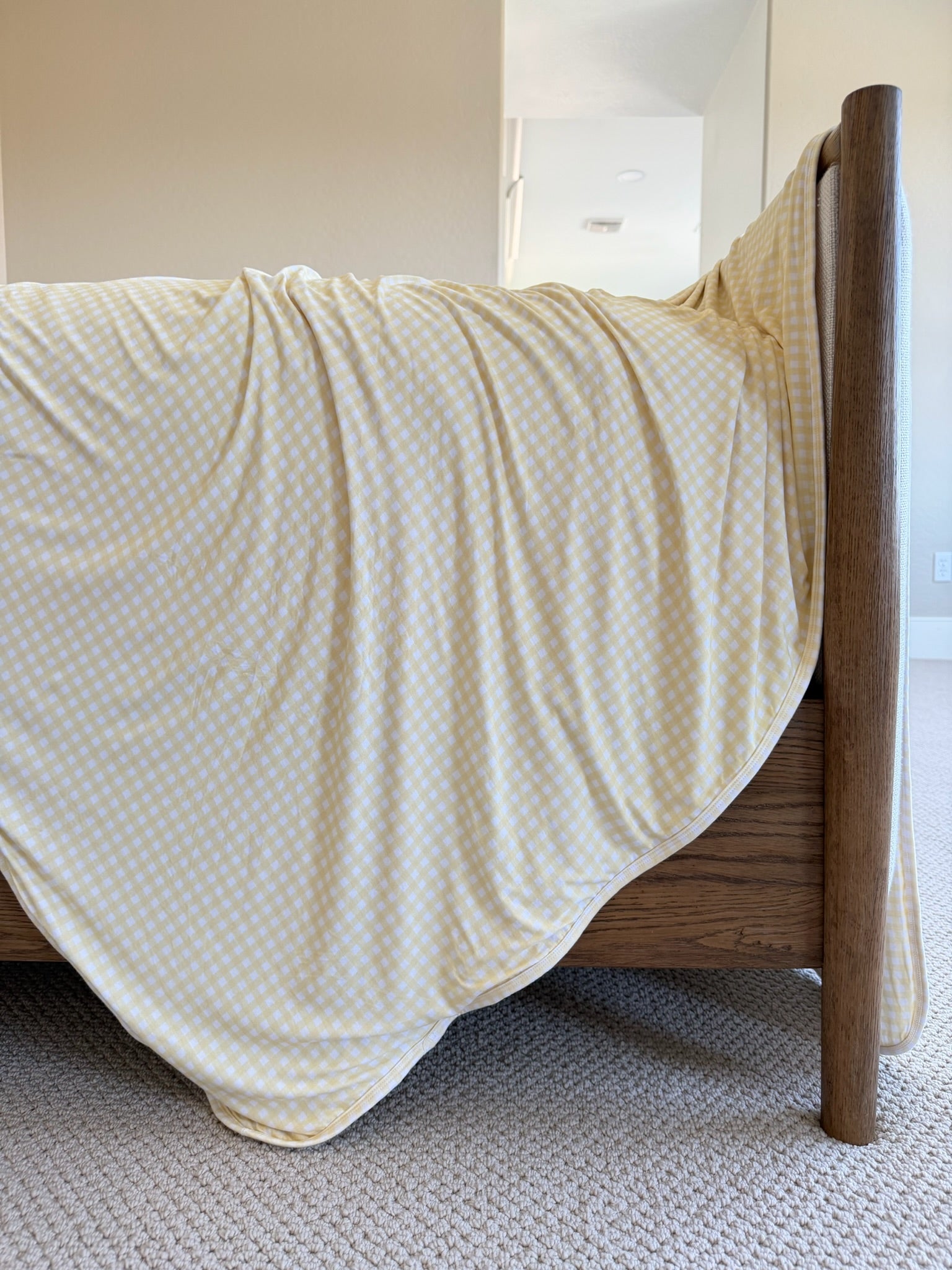 A "sunshine" yellow gingham blanket drapes over the corner of a wooden bed frame in a bright, carpeted bedroom with light-colored walls.