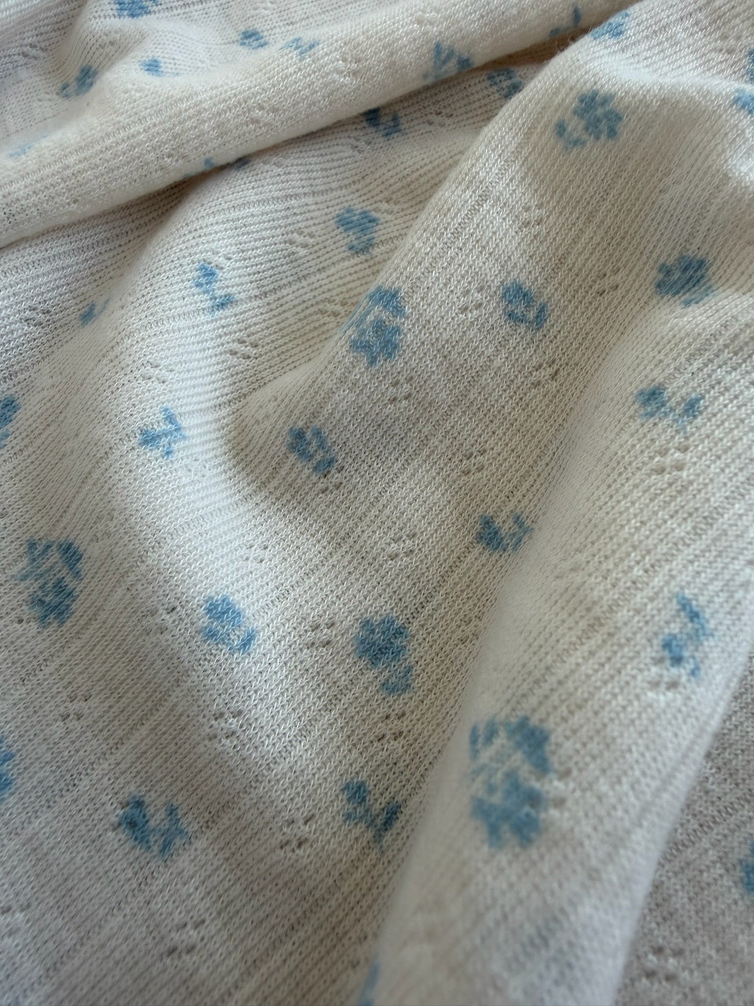 A close-up of the blue willow / pointelle fabric, featuring soft, cream-colored ribbed material adorned with delicate blue floral patterns. The gently folded, textured surface resembles a luxurious blanket.