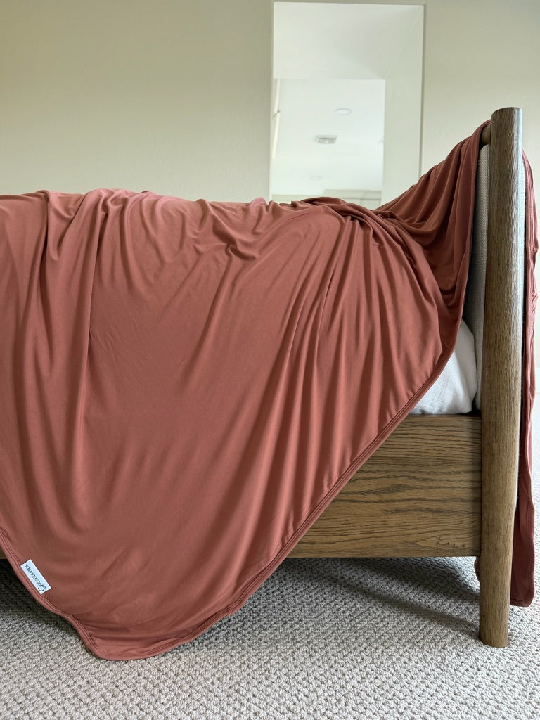 A luxurious Serenity blanket in a rust hue drapes over the side of a wooden bed frame on a light carpeted floor in a neutral-toned room.