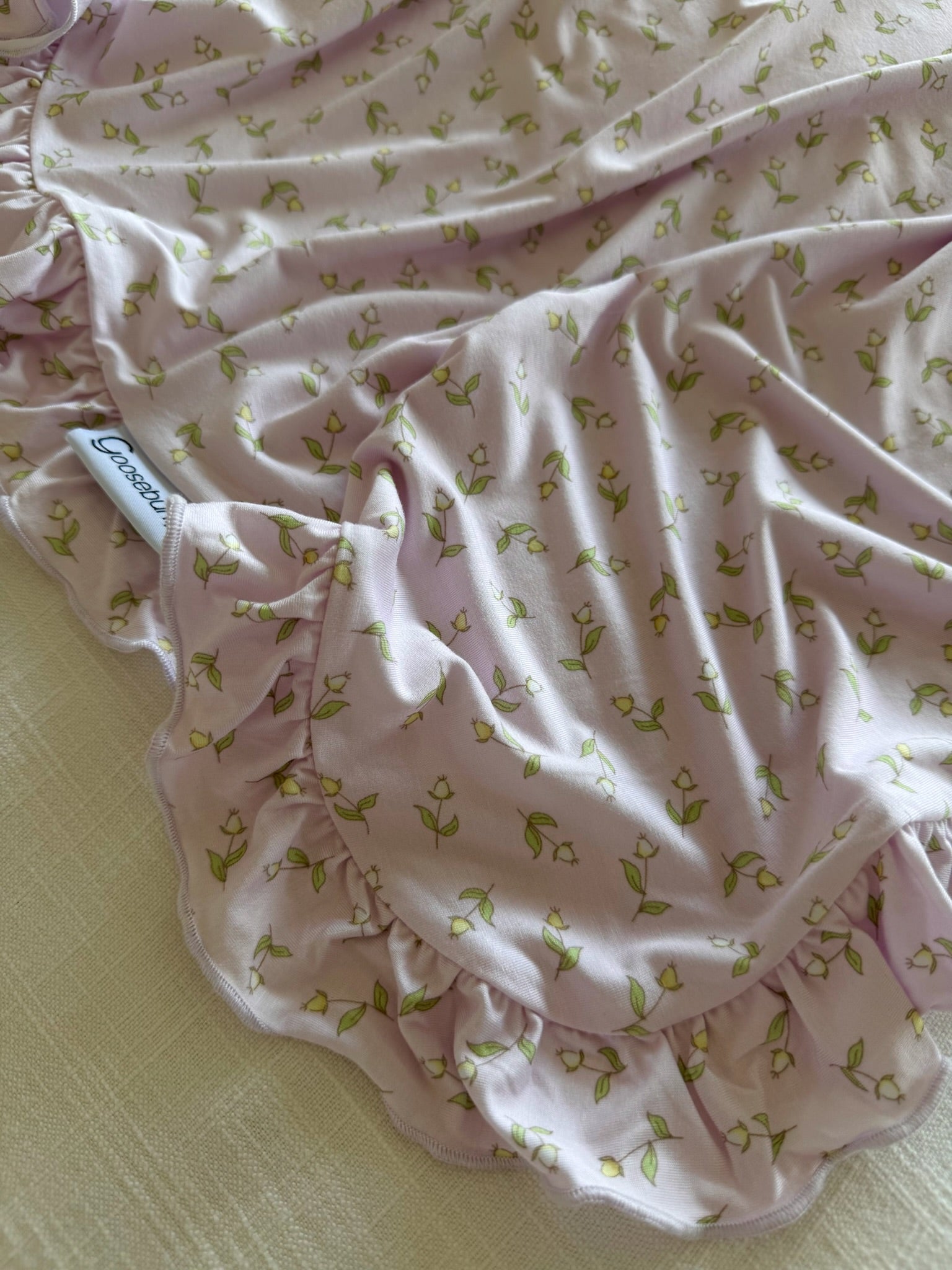 A close-up of the rosebud / ruffle light pink bamboo blanket with green and yellow floral pattern, ruffled edges, and a white tag in cursive. The luxurious blanket is slightly wrinkled and lies on a textured surface.