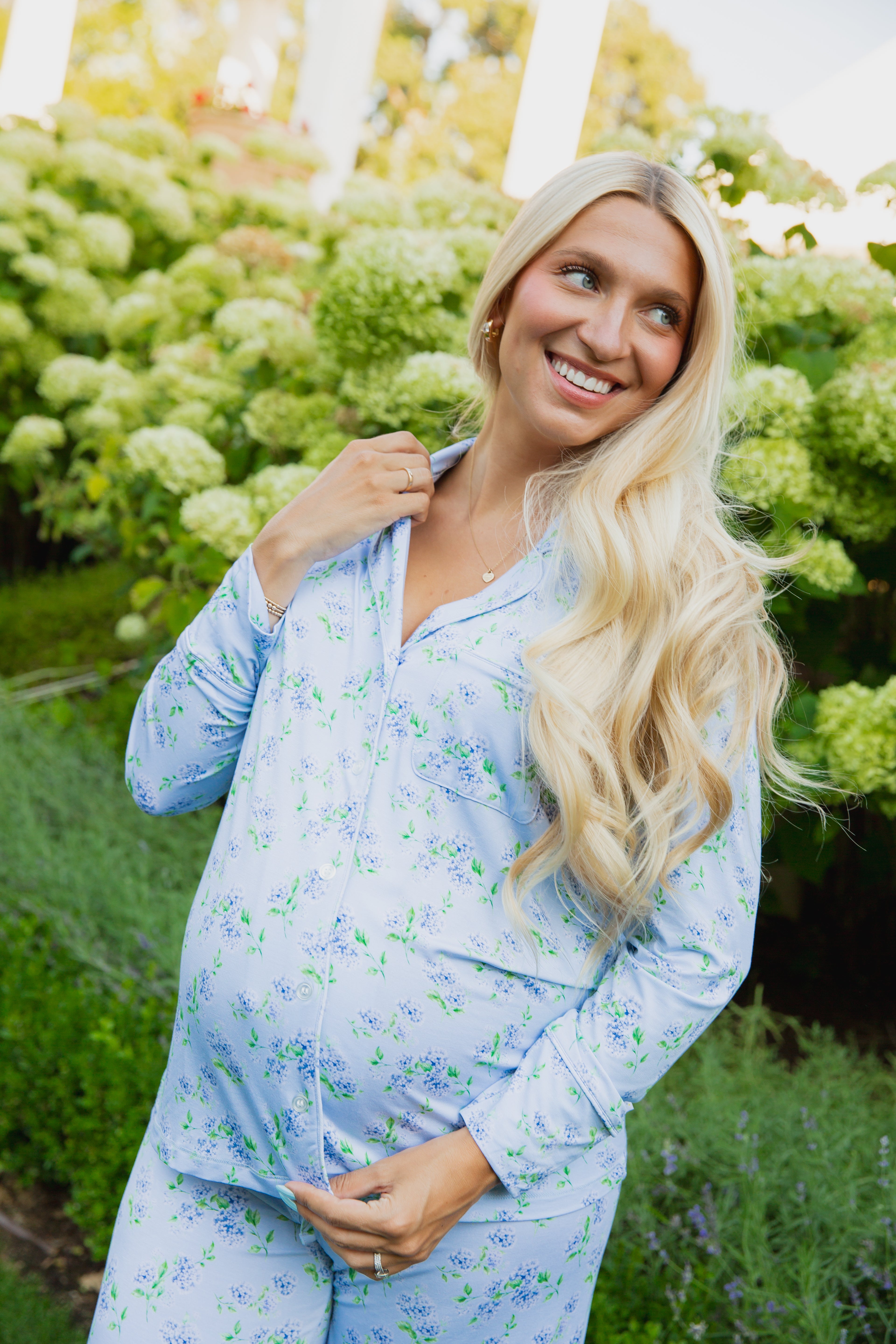A pregnant woman with long blonde hair, wearing the hydrangea pajama set (long pant) from the Sarah Sweeten Goosebumps Collection in a classic hydrangea print, smiles while standing in a lush garden with green hydrangea bushes in the background.