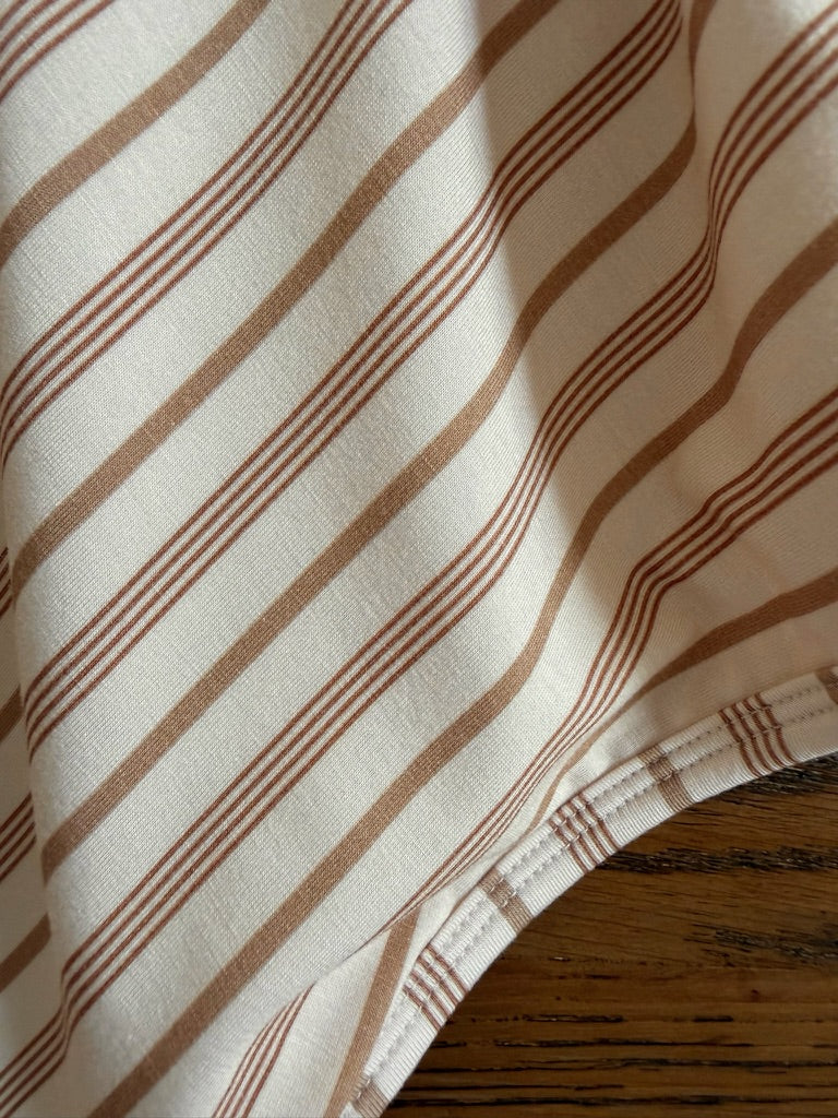 A close-up of the toast and honey travel blanket in beige, featuring diagonal brown stripes, draped elegantly over a wooden surface. The edge of this luxurious blanket, complete with visible stitching, is positioned at the lower right corner.