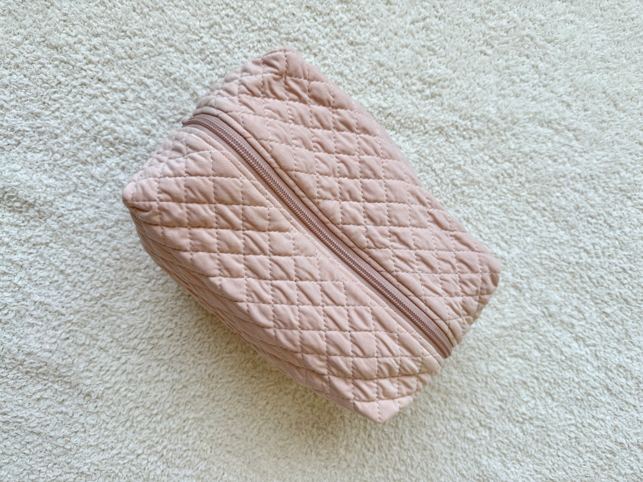 The whisp / travel bag, a pink quilted rectangular pouch from the Lisa Wilson Goosebumps Collection, rests on a light textured surface—perfect for carrying your essentials on the go.