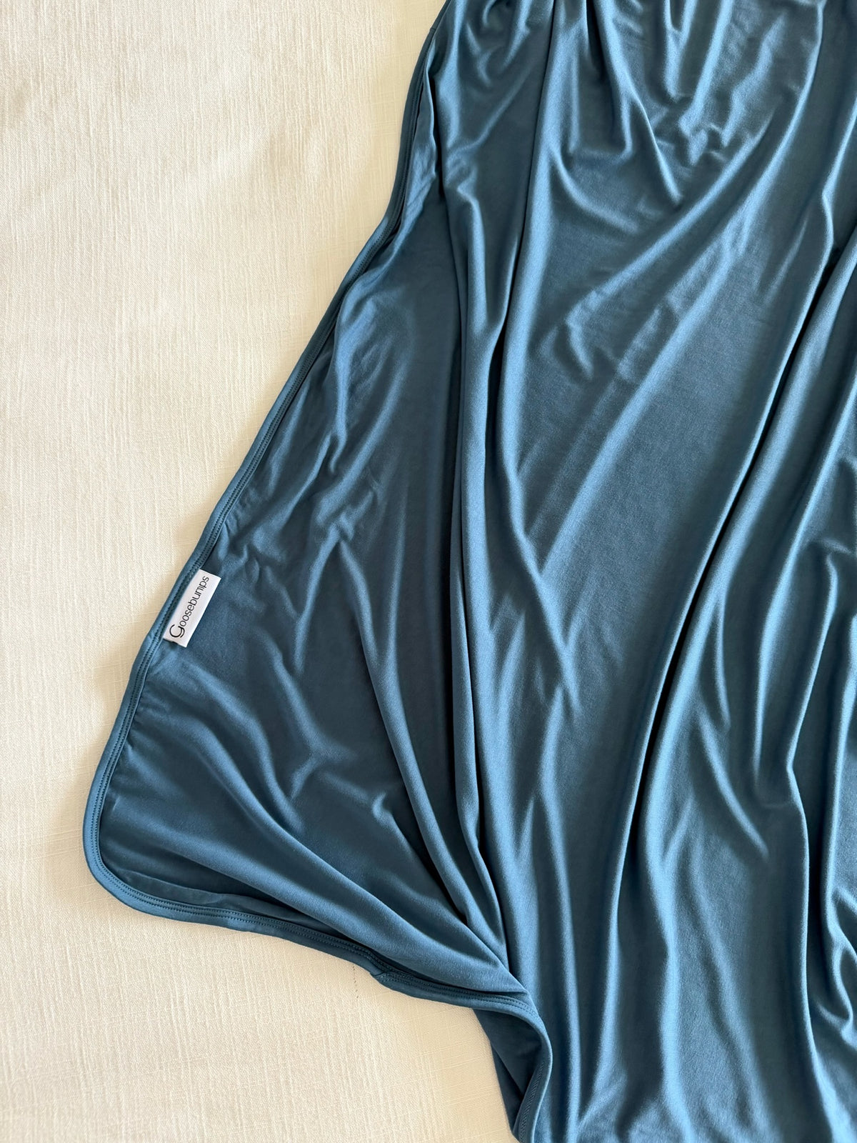 The “tidal wave” blanket, in a silky blue fabric with a small white tag on the edge, lies partially spread out on a light-colored textured surface.