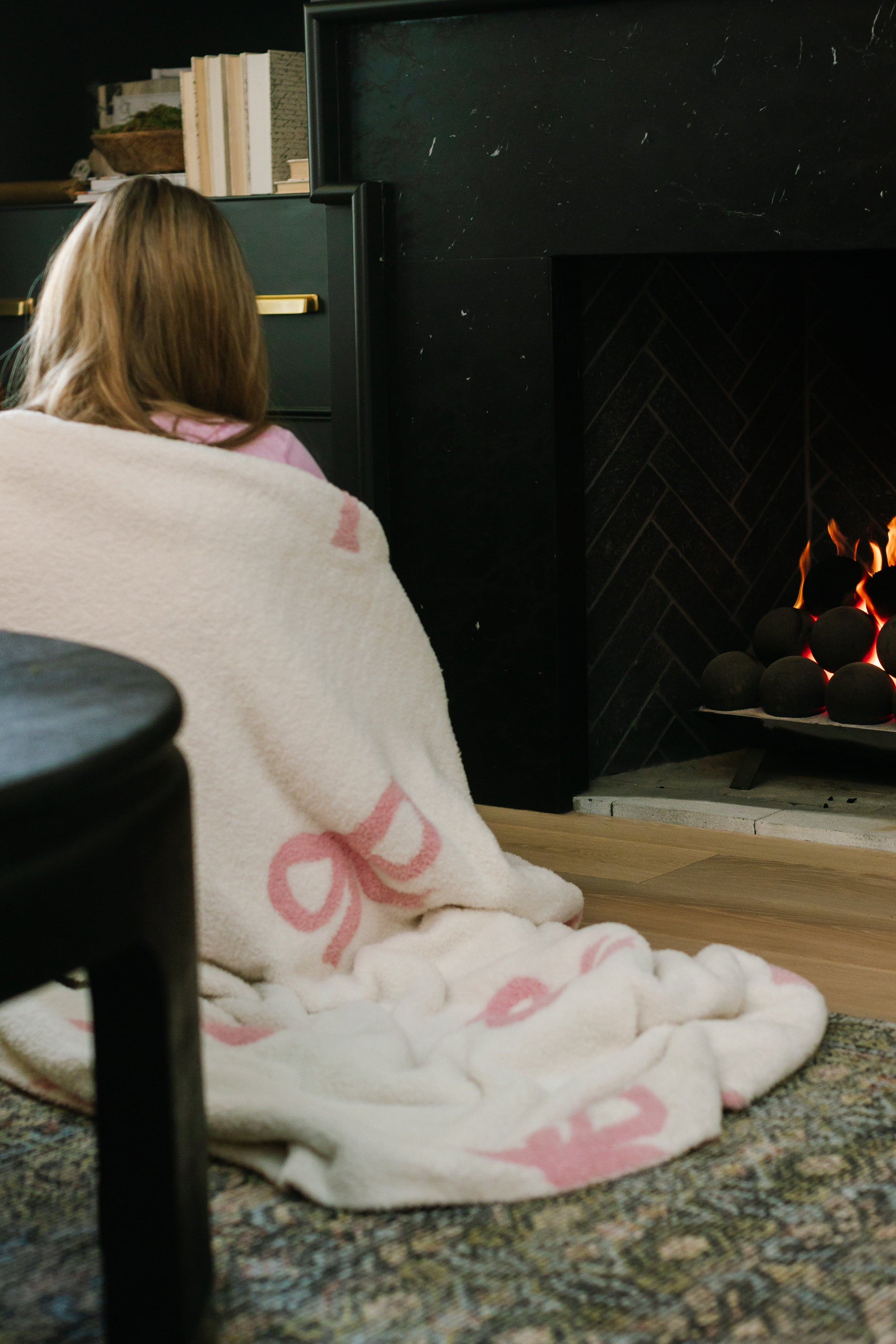 Wrapped in the bowie / pink / plush blanket, a person with long hair sits on a rug near a glowing fireplace in a cozy living room, with shelves of books in the background.