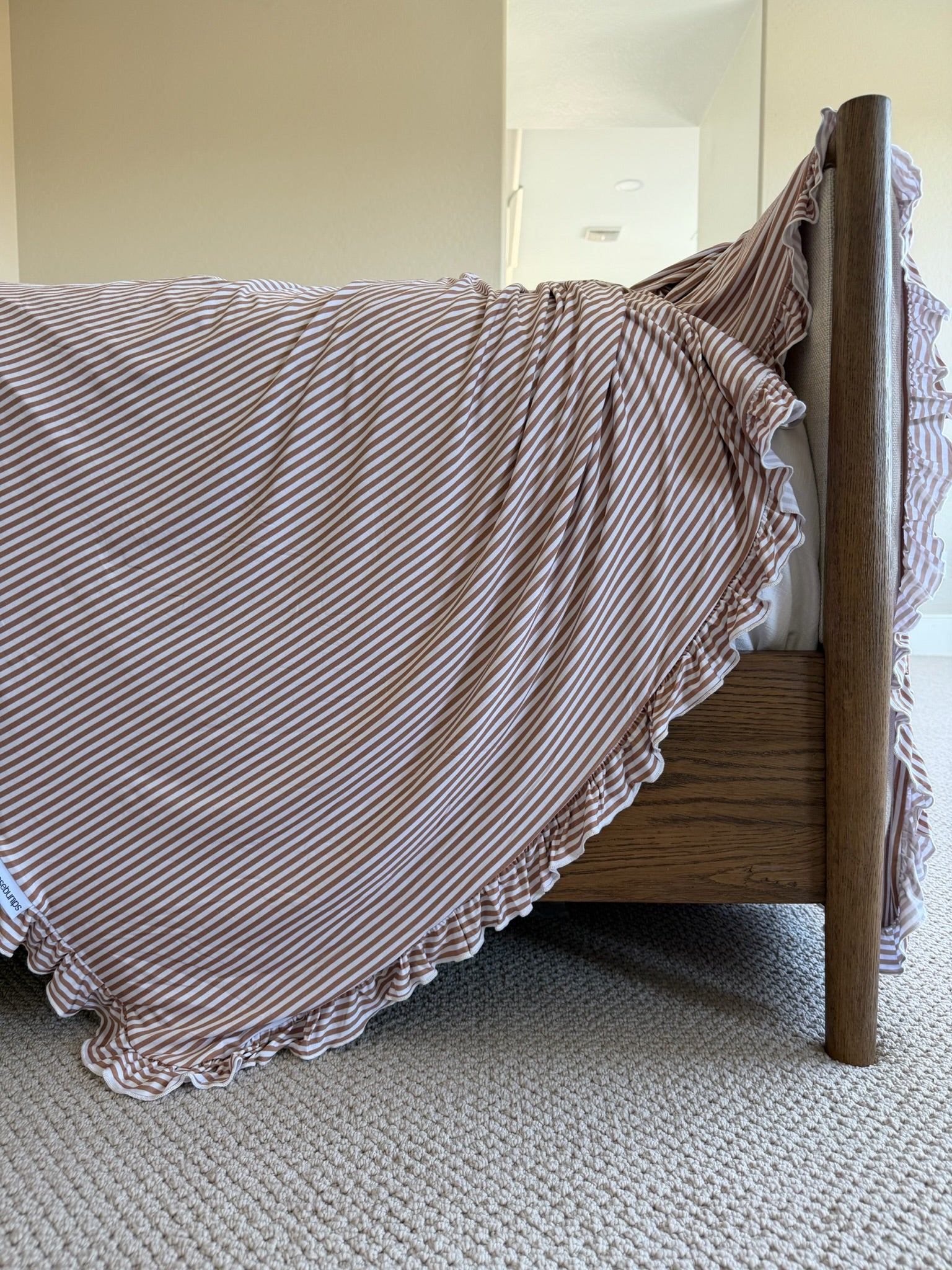 A close-up of a bed with a wooden frame, featuring the drift / ruffle adult swaddle in light brown and white stripes with ruffle trim, draped over the side and touching a beige carpeted floor.