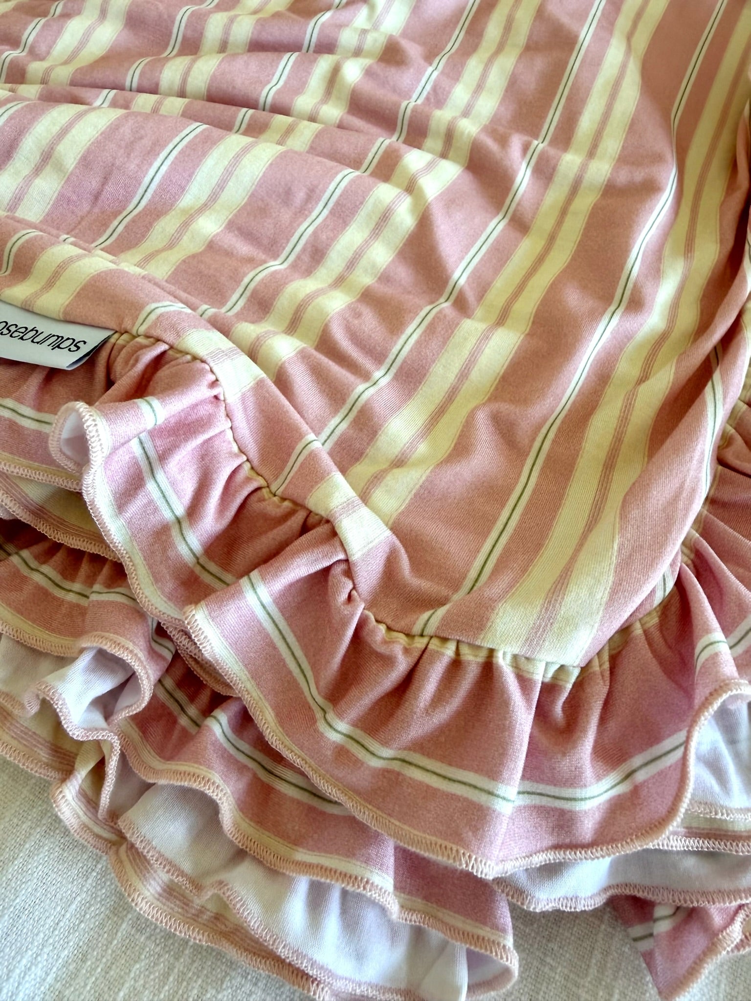 A close-up of the sherbet / ruffle blanket, featuring pink and white horizontal stripes with ruffled edges, draped in soft folds on a light-colored surface.