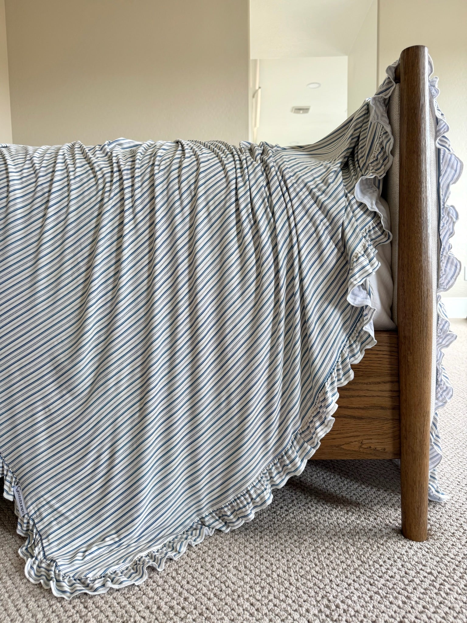 The beach house / ruffle features a luxurious, light blue and white striped blanket with ruffled edges draped over a wooden footboard, cascading onto a beige carpeted floor in a softly lit room.