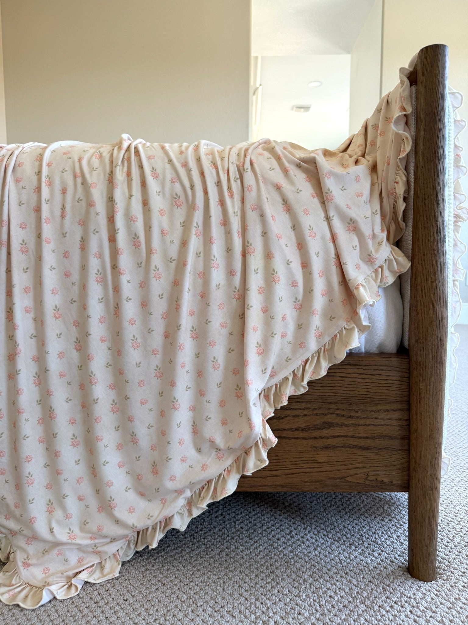 The petal / ruffle wooden bed features a light cream, luxuriously soft blanket with small pink floral prints and ruffled edges, gracefully draping over the side in a brightly lit, cozy room.