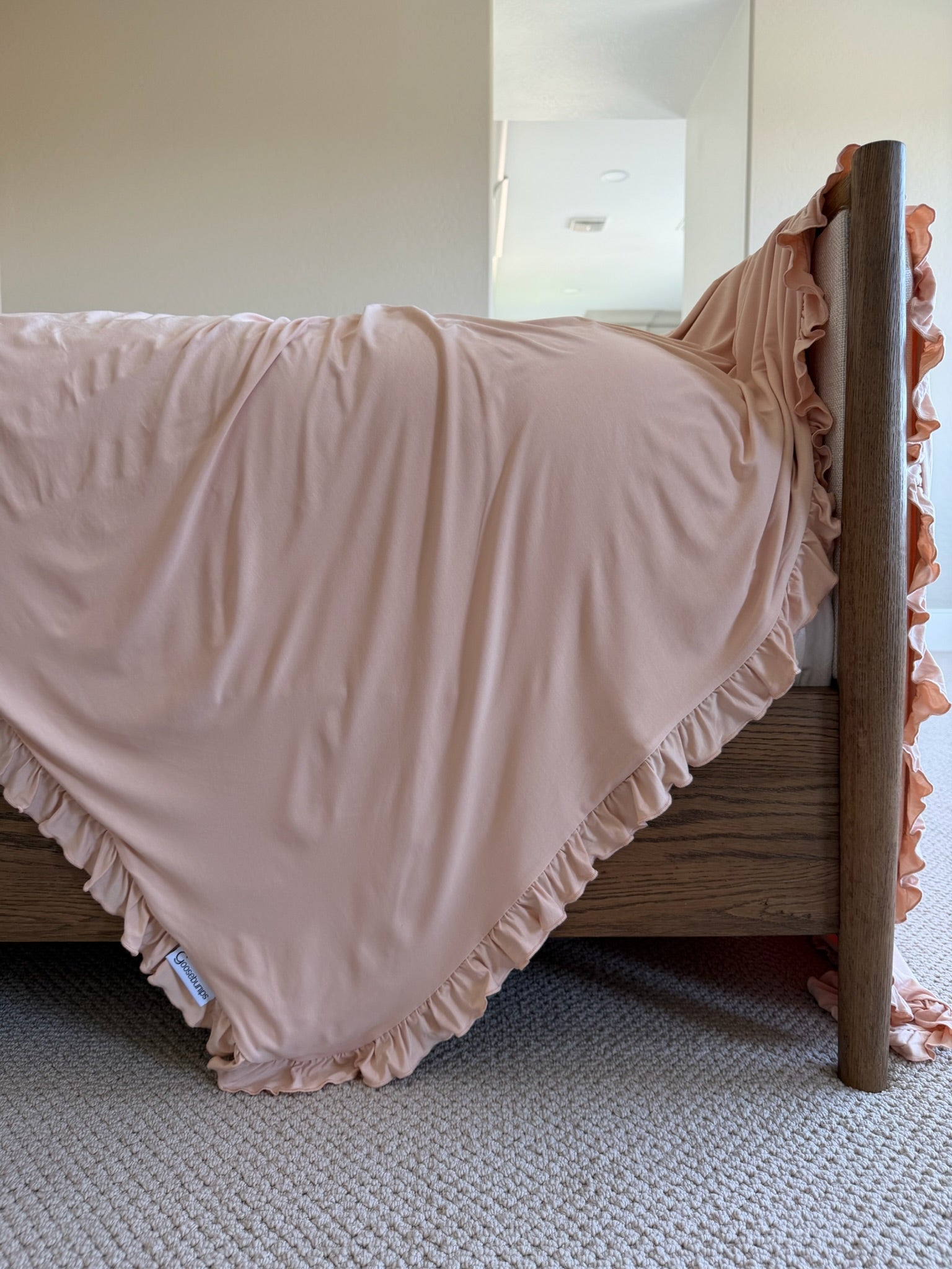 The whisp / ruffle blush pink blanket drapes over a wooden bed frame, its ruffled edge just touching the beige carpet in a bright, neutral bedroom.