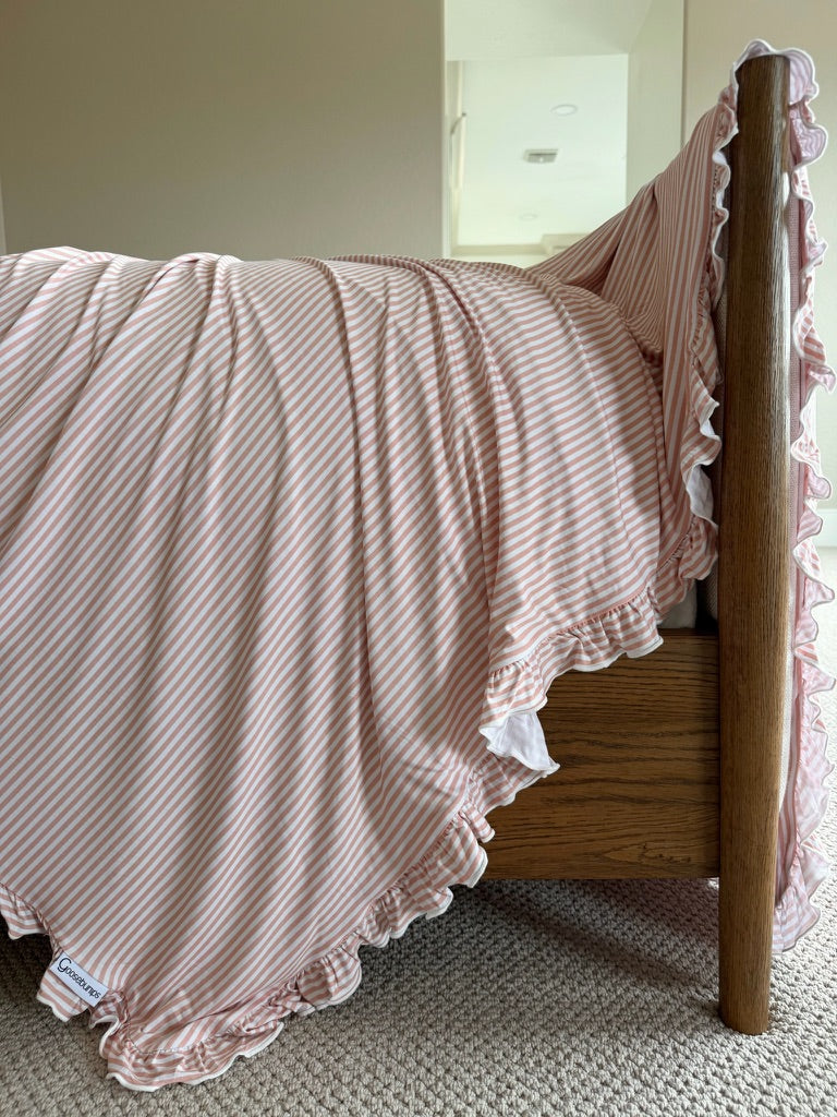 A heritage / ruffle blanket in pink and ivory stripes, featuring elegant ruffled edges, is draped over the side of a wooden bedframe, its luxurious fabric standing out softly against the beige carpeted floor below.
