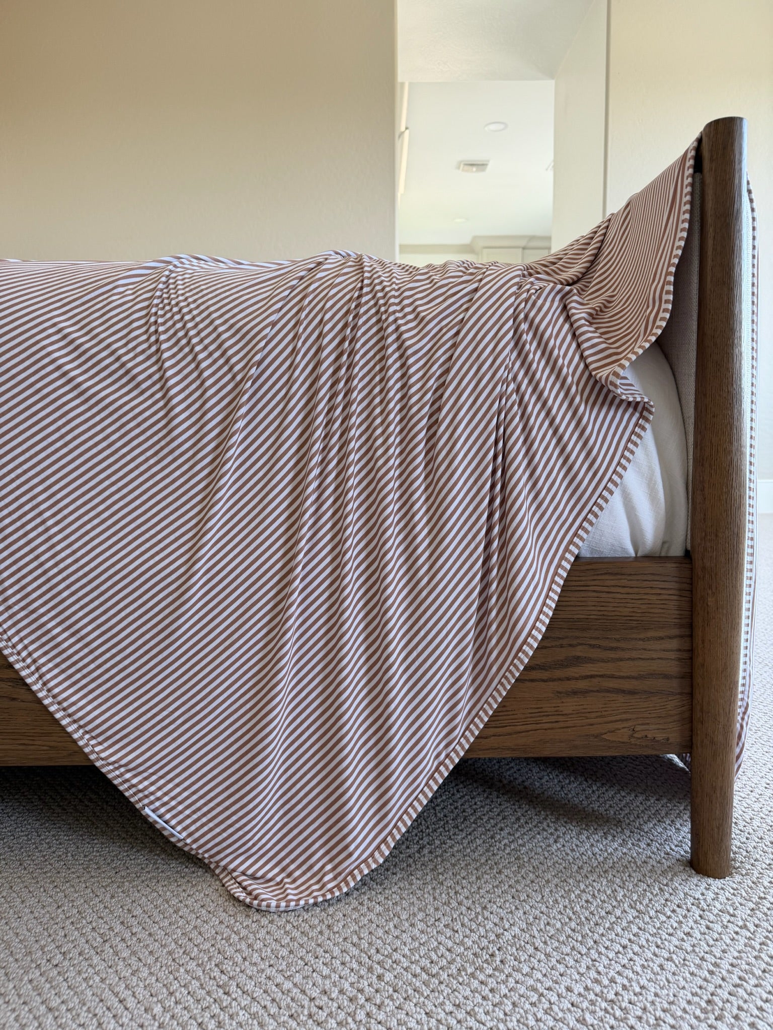 A bed with a wooden frame is covered by the drift white and light brown striped blanket, which drapes over the side and touches the beige carpeted floor in a softly lit room.