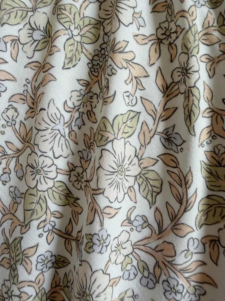 Close-up of heirloom / ruffle fabric with a floral pattern in soft beige, green, and white tones. The slightly wrinkled material features detailed outlines of leaves and flowers throughout the design, adding delicate texture to this elegant floral blanket.