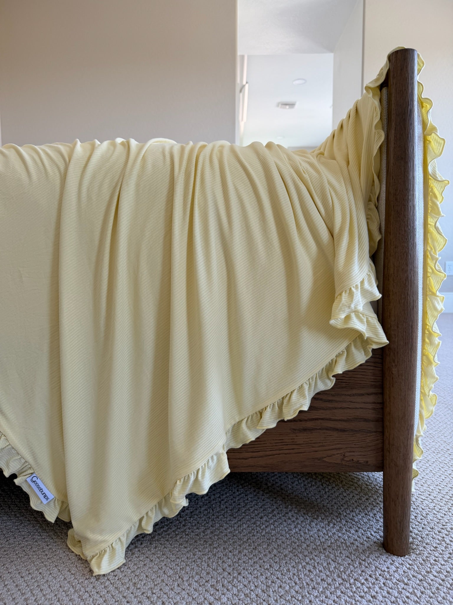 A butter yellow ruffle blanket is draped over a wooden bed frame, set against beige carpet and a softly lit hallway in the background.
