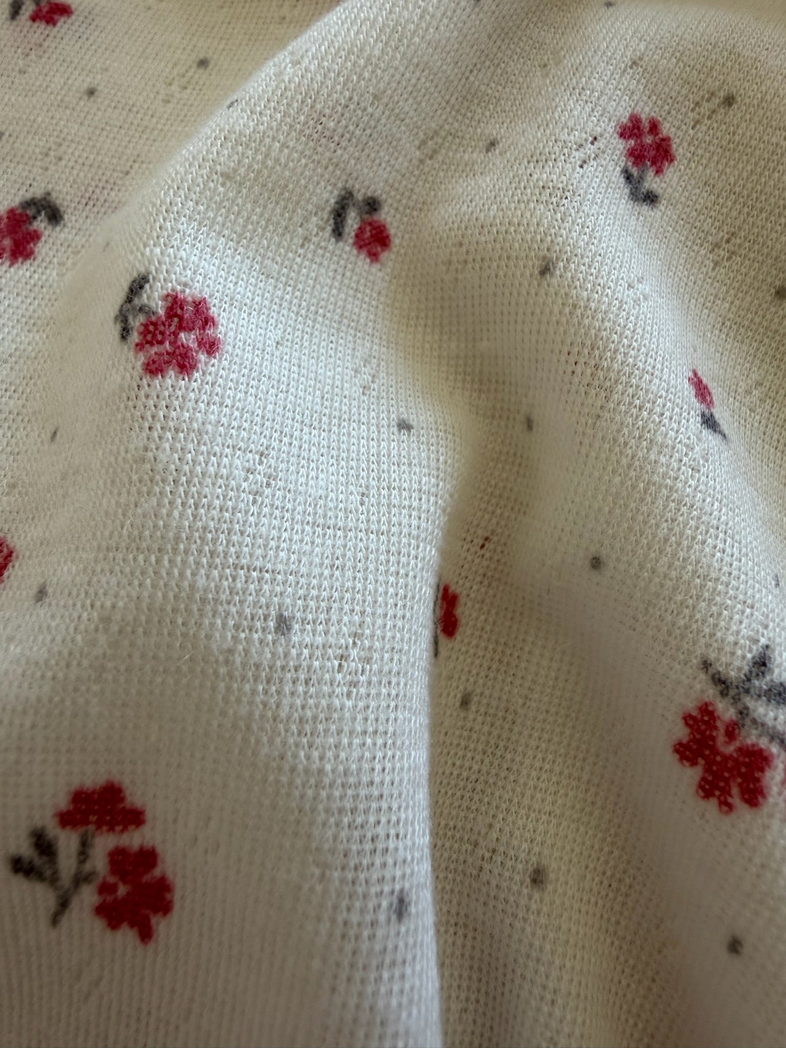 A close-up of ivory bloom / pointelle fabric shows its ribbed texture, red and black floral prints, and visible pilling, highlighting the worn look of this modern blanket.