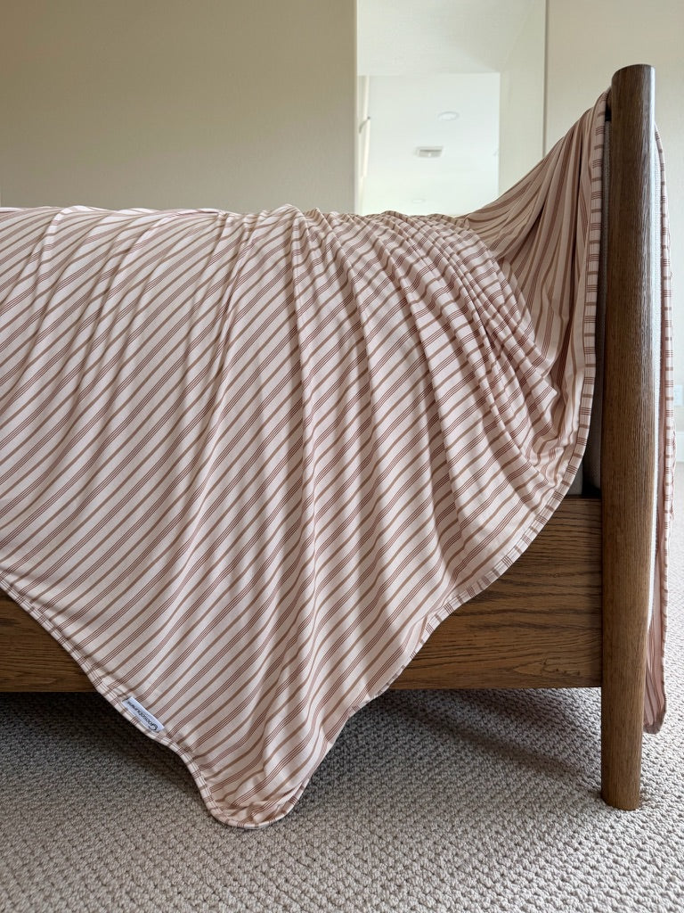 A “toast and honey” bamboo blanket, featuring luxurious beige and white stripes, is draped over the corner of a wooden bed frame and rests on a light beige carpet in a softly lit bedroom.