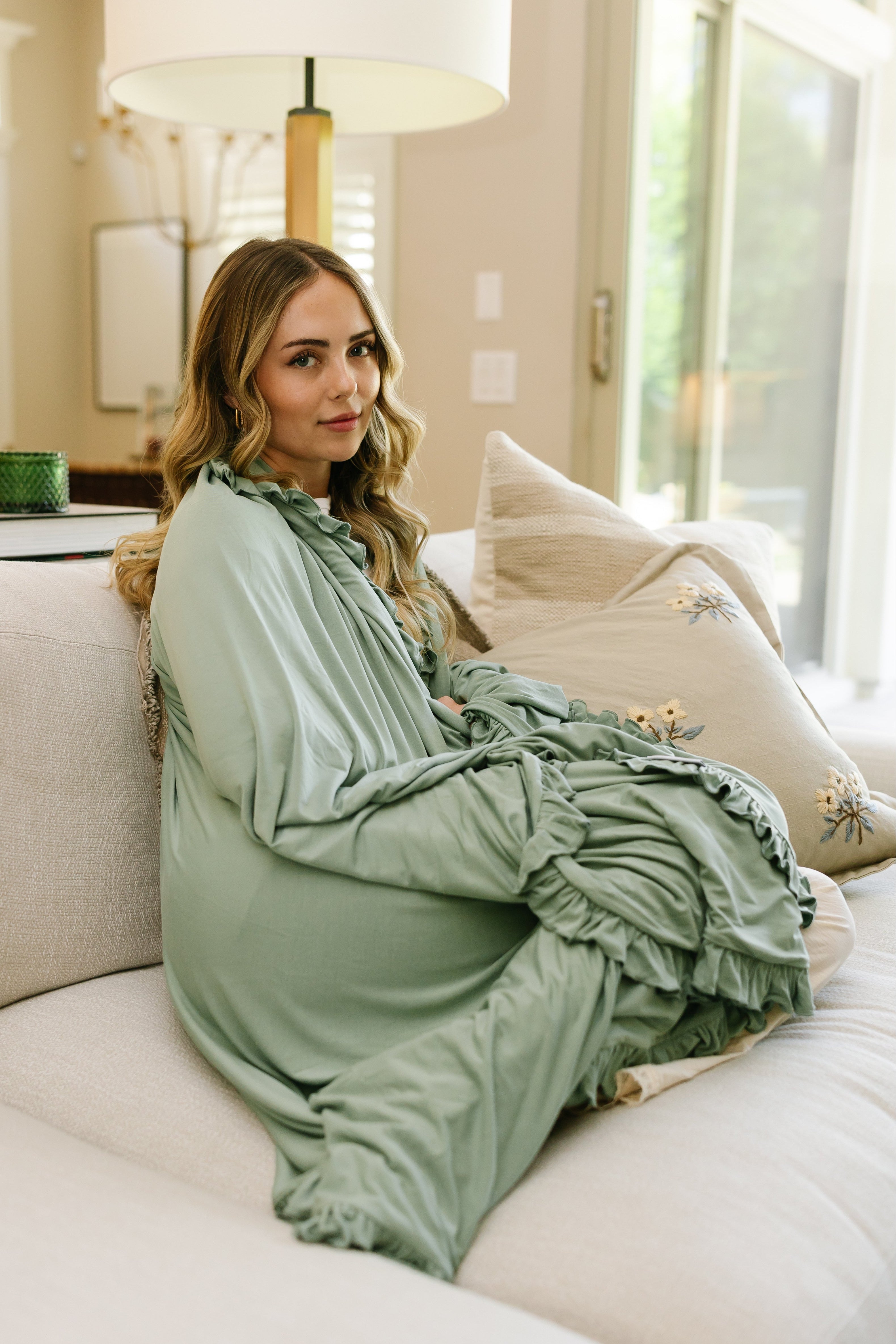 A woman with long, wavy blonde hair sits on a light sofa in a bright, modern living room, draped in a basil / ruffle blanket over her flowing sage green dress, looking at the camera with a neutral expression.