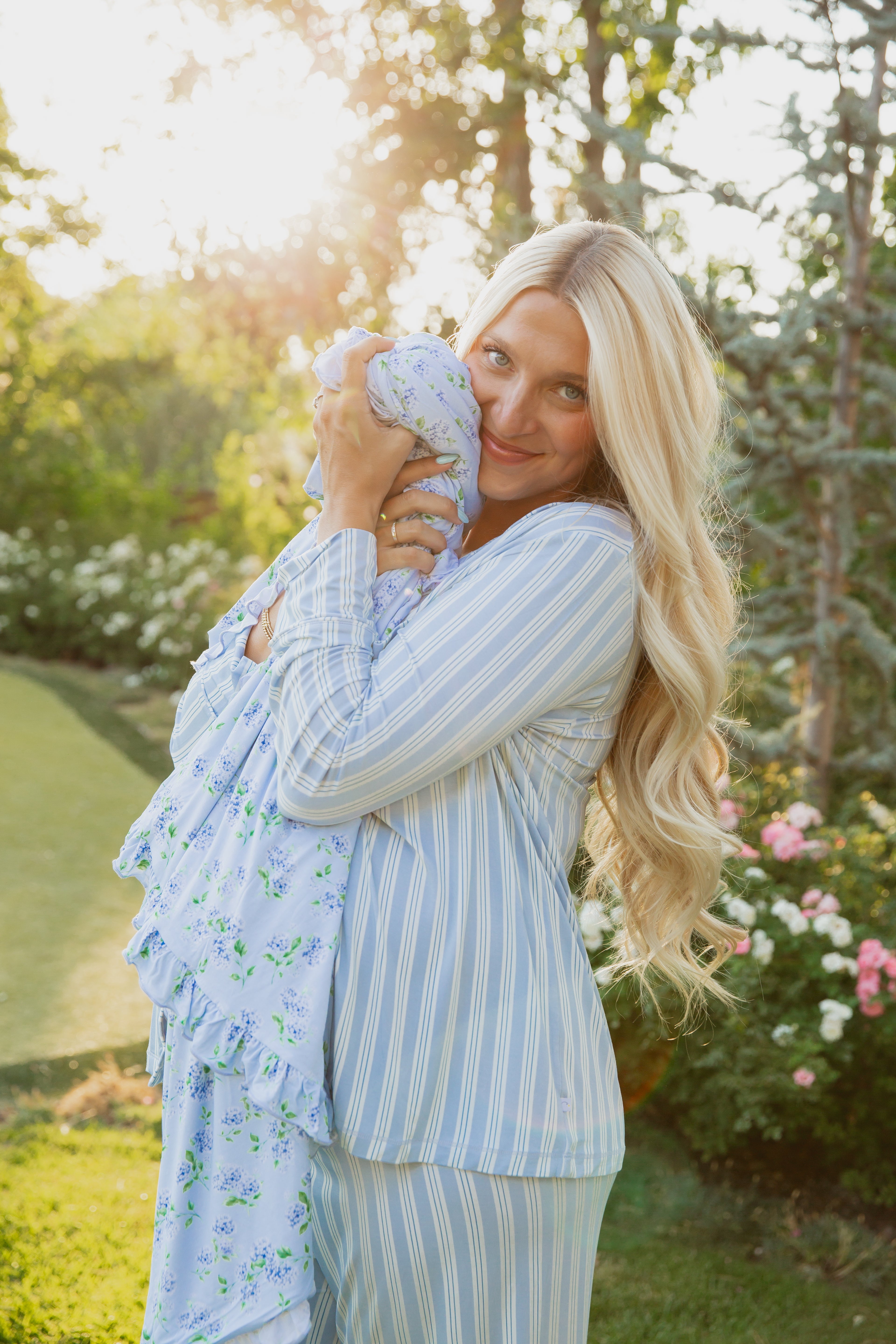 A woman with long blonde hair, dressed in blue striped pajamas, lovingly cradles a baby wrapped in the hydrangea / ruffle bamboo blanket as they enjoy a sunlit garden.