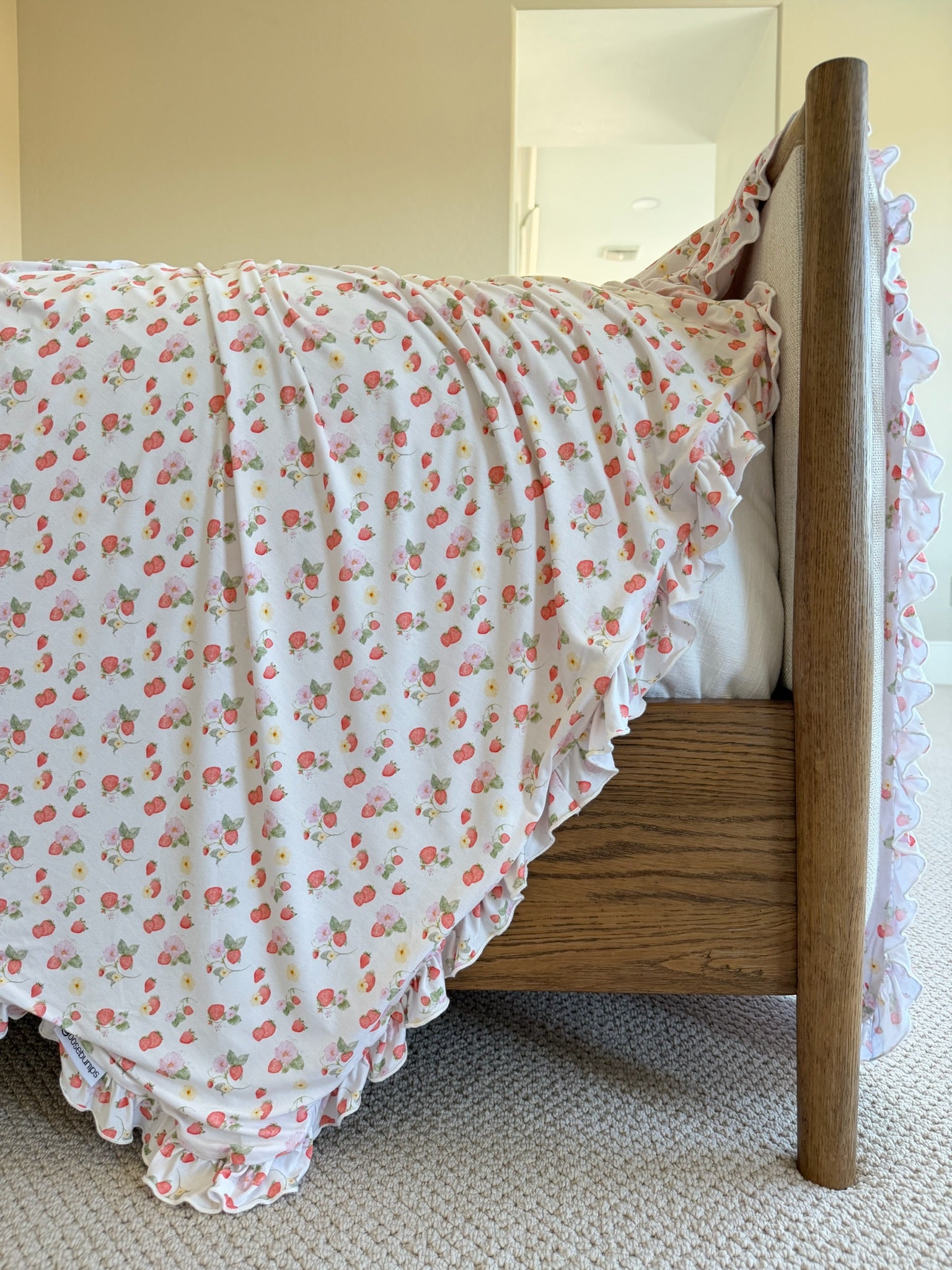 A wooden bed is draped with the freshly picked / ruffle blanket, featuring small pink strawberries and ruffled edges. The room has beige walls and soft, carpeted flooring.