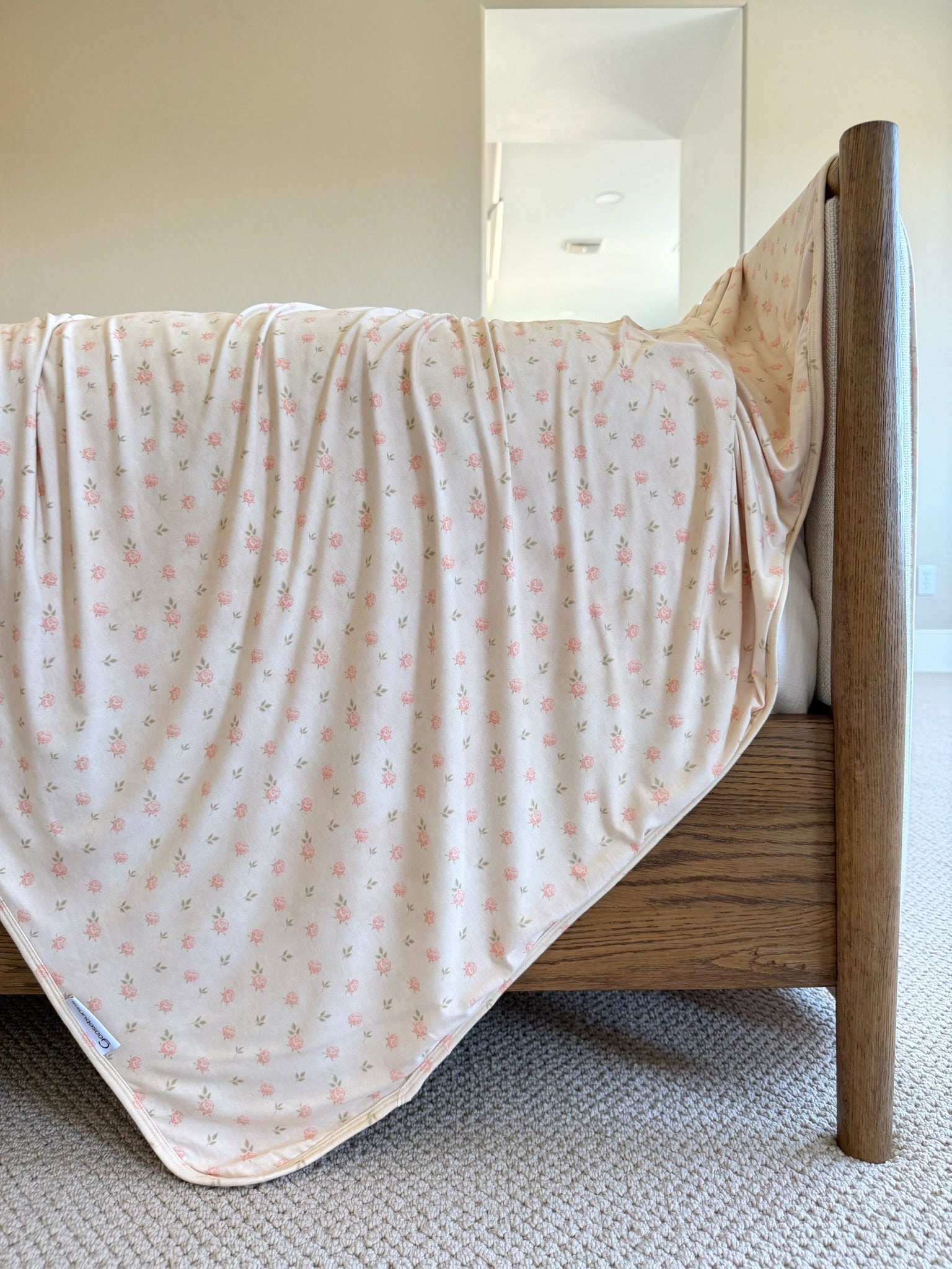 A pale pink bamboo blanket, petal, with a small floral pattern is draped over the side of a wooden bed on a beige carpet in a bright, minimalist bedroom.