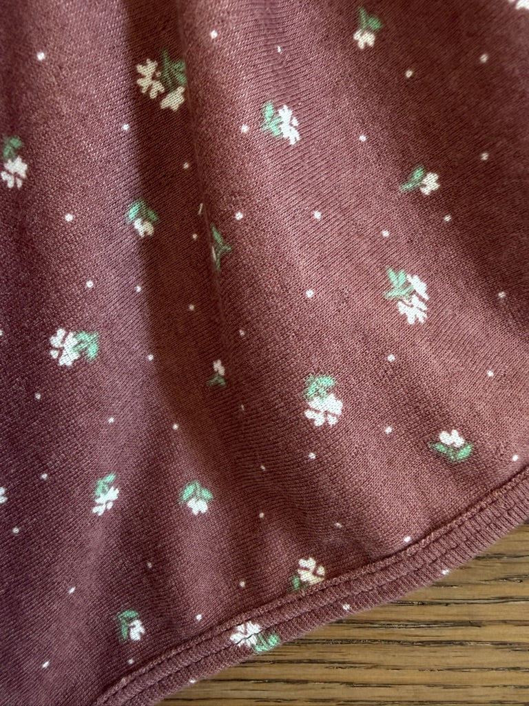 A close-up of botanical / pointelle: maroon ribbed fabric with a small white and green floral pattern and tiny white dots, draped over wood.
