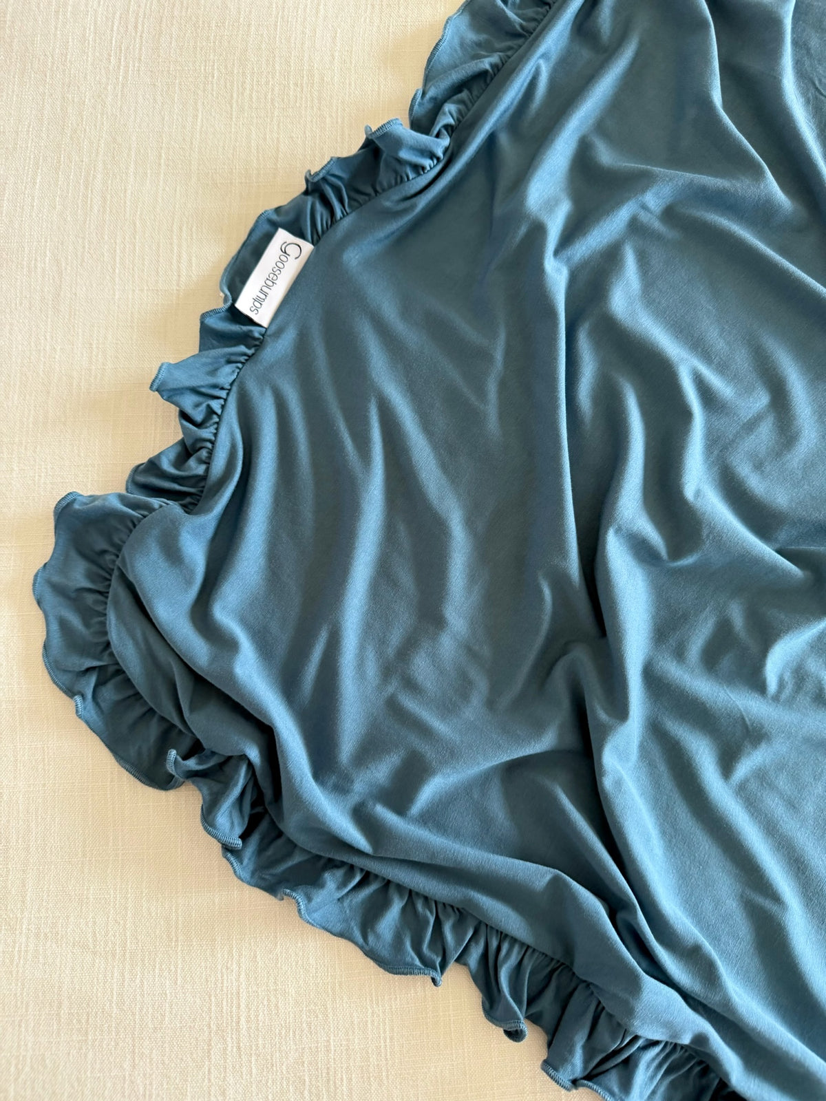 A tidal wave / ruffle blanket in dark teal, featuring luxurious ruffles and a Goosebumps brand tag, draped over a light-colored, textured surface.