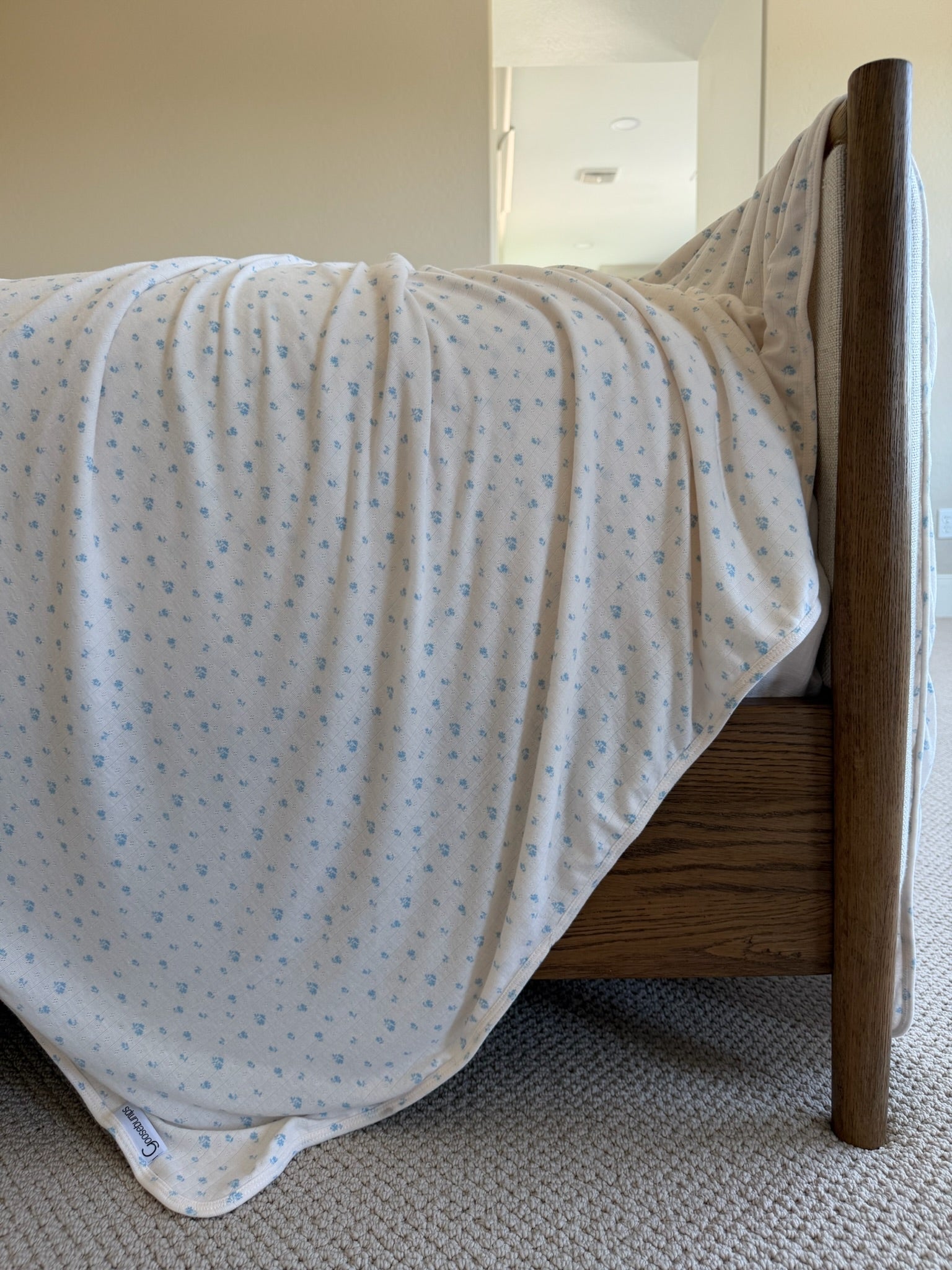 The blue willow / pointelle blanket, featuring cream-colored ribbed fabric and small blue floral patterns, is draped over a wooden bed frame in a carpeted room with neutral walls.