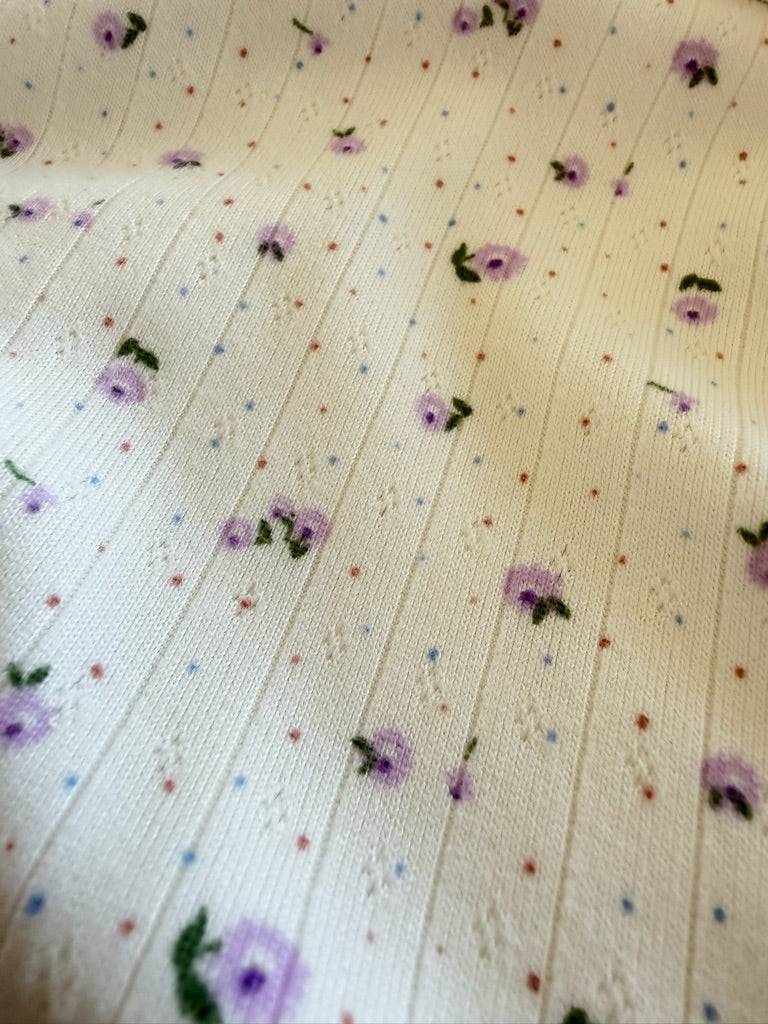 Close-up of iris / pointelle: a cream-colored ribbed fabric with delicate lavender flowers and scattered red and blue dots across its textured surface.