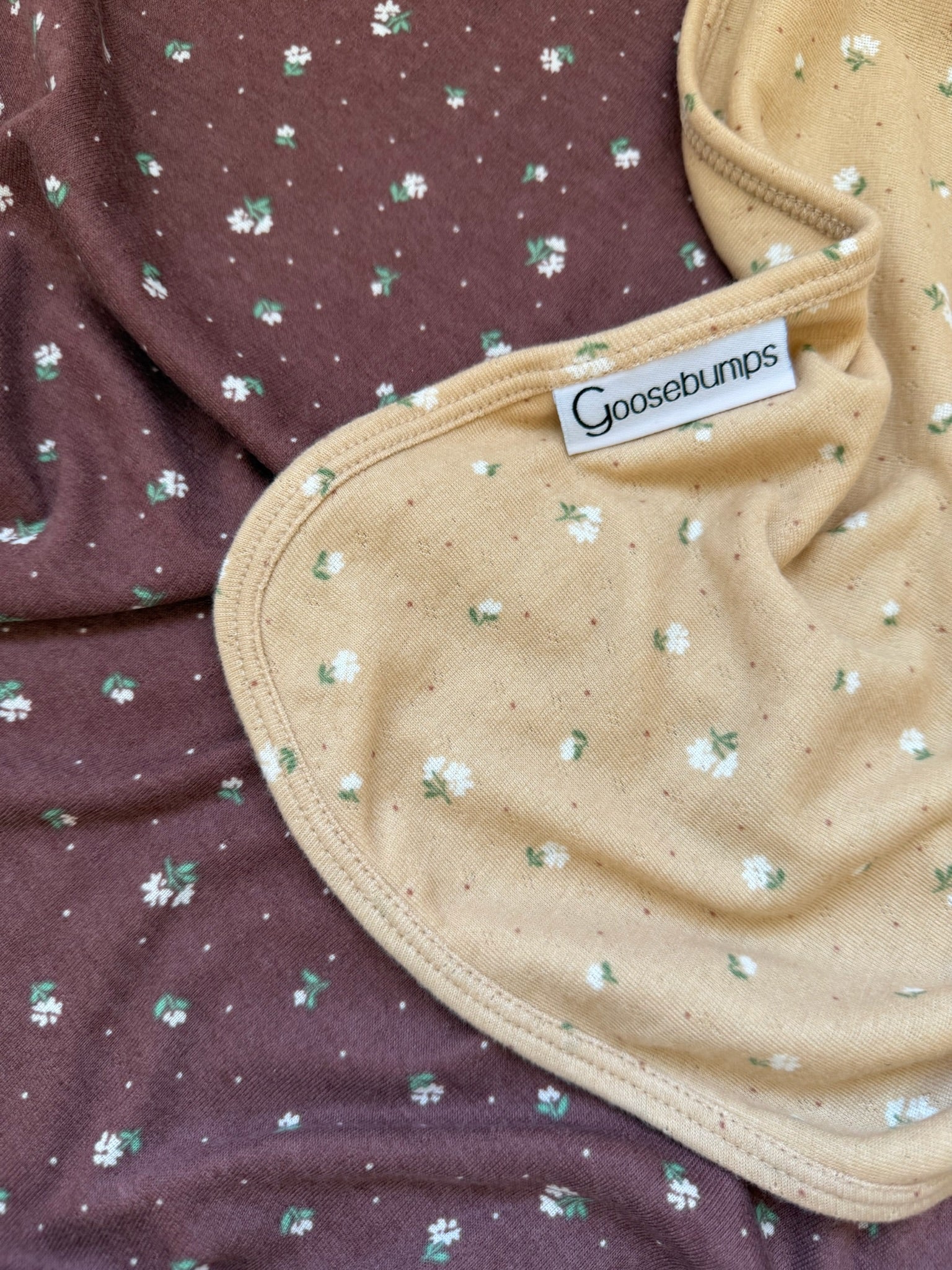 A close-up of two fabrics from the botanical / pointelle collection: one brown with small white and green florals, and one beige ribbed pointelle fabric with similar floral patterns and a Goosebumps label attached.