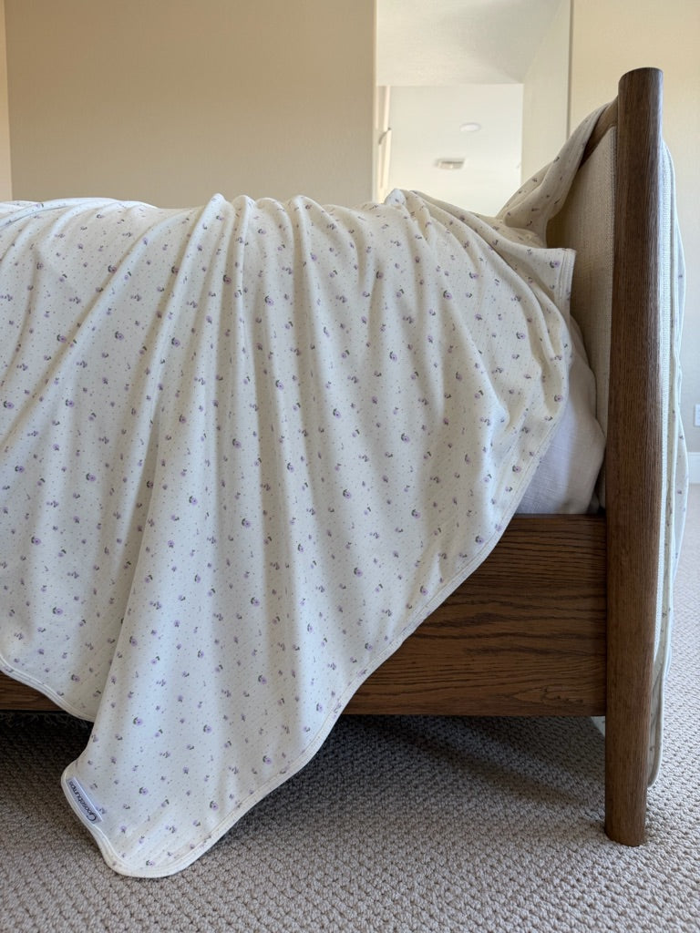 A close-up of a wooden bed with the iris / pointelle blanket in cream, decorated with small lavender flowers, draped over the side. The beige carpet and softly lit, neutral room create a serene backdrop.