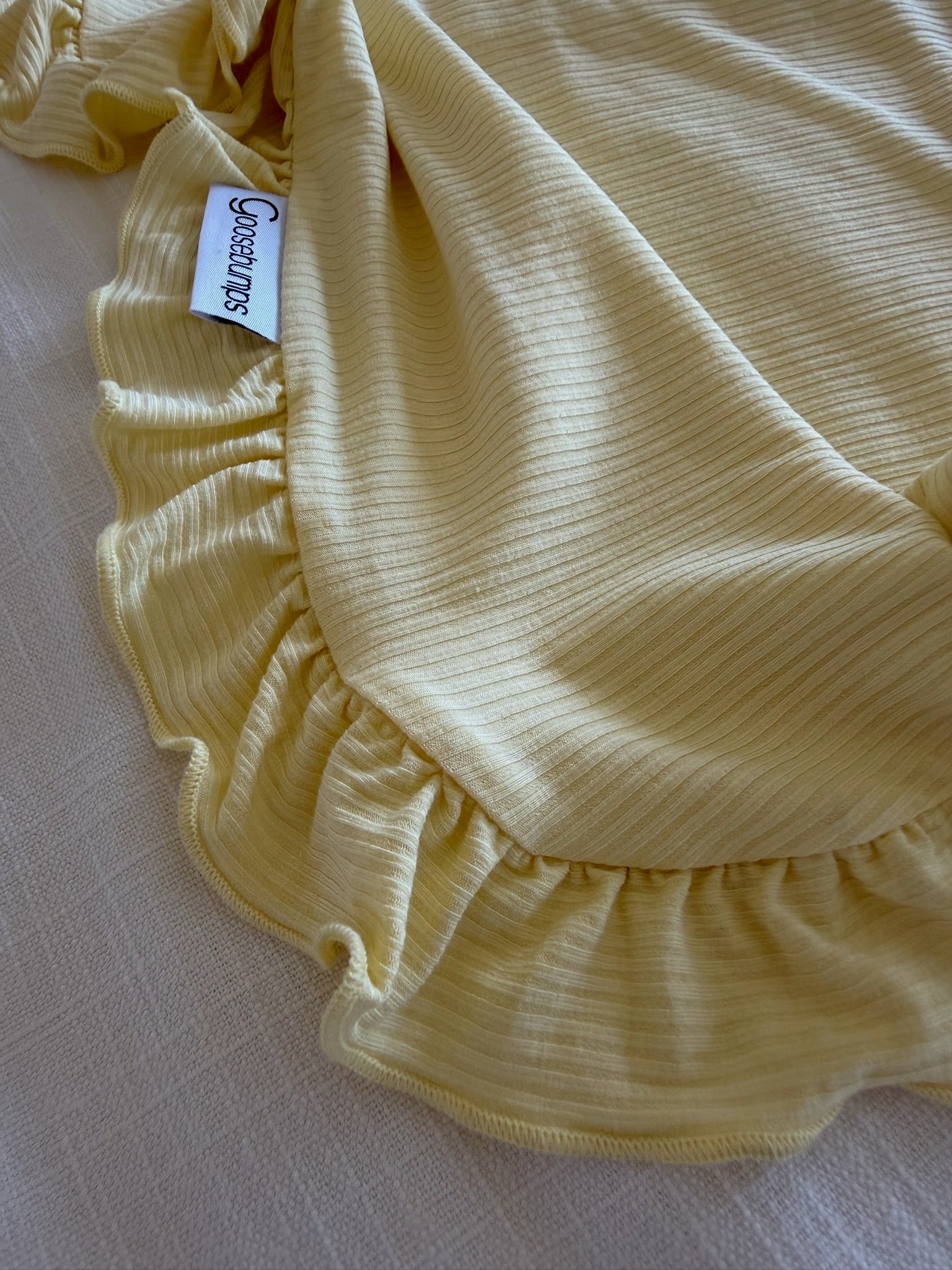 A close-up of the butter / ruffle bamboo blanket, featuring soft yellow ribbed fabric and ruffled edges, rests on a light textured surface. The visible “Generous” tag highlights this luxurious blanket’s unique design.