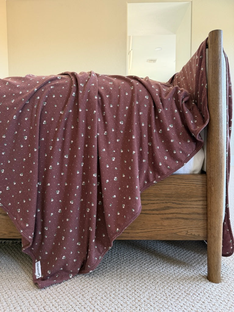 A luxurious maroon botanical / pointelle blanket with small white floral patterns is draped over the edge of a wooden bed frame, resting on a textured beige carpet in a bright, softly lit room.