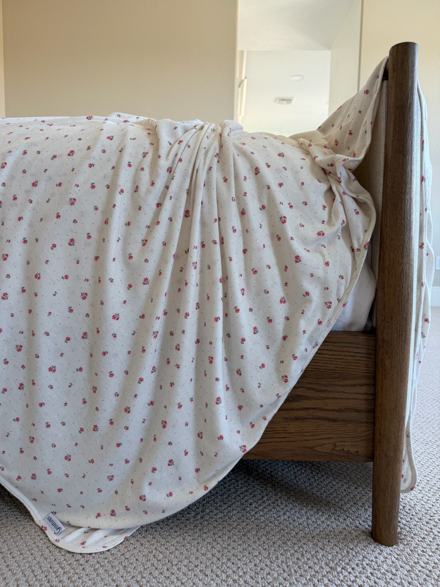 The ivory bloom / pointelle blanket, featuring small red floral patterns, is draped loosely over a wooden footboard in a carpeted room with beige walls.