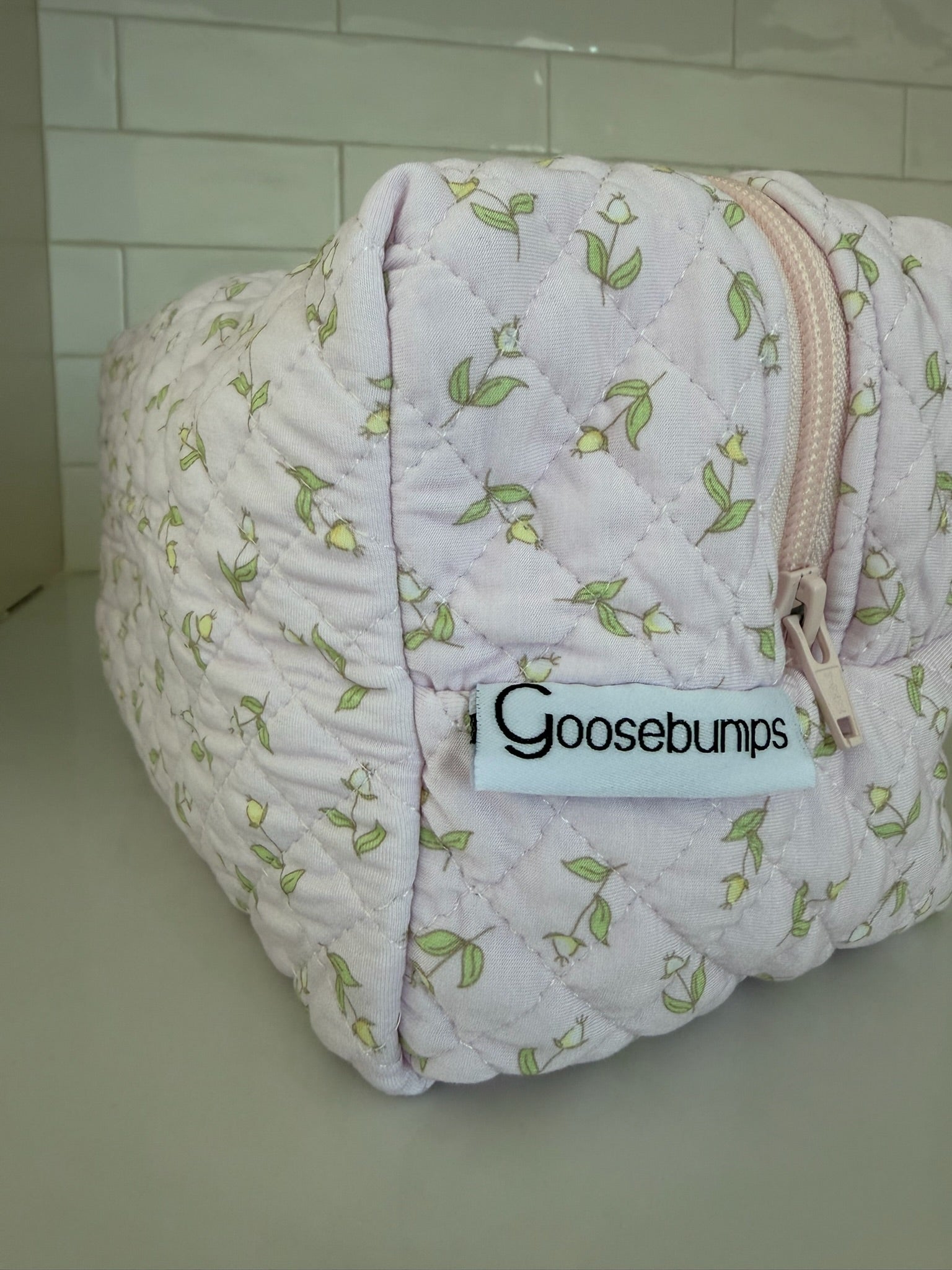 A quilted pastel pouch featuring a delightful floral pattern, beige zipper, and a label that reads "rosebud / travel bag," displayed on a white surface with a tiled background—perfect for adding a touch of style to your travel accessories collection.
