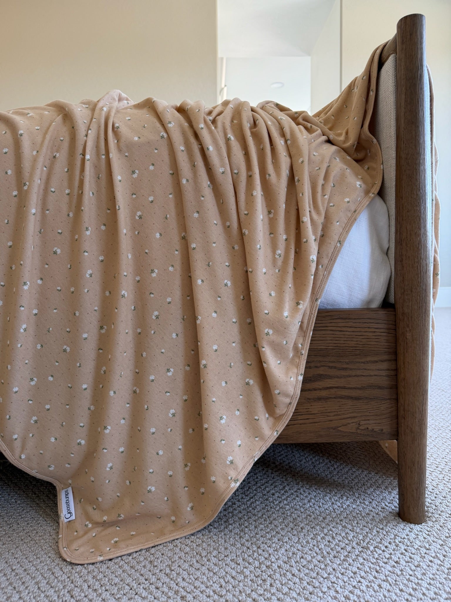 The iced coffee / pointelle blanket, a light brown luxurious ribbed pointelle fabric with small white floral patterns, is draped over a wooden bed frame on a beige carpet in a softly lit bedroom.