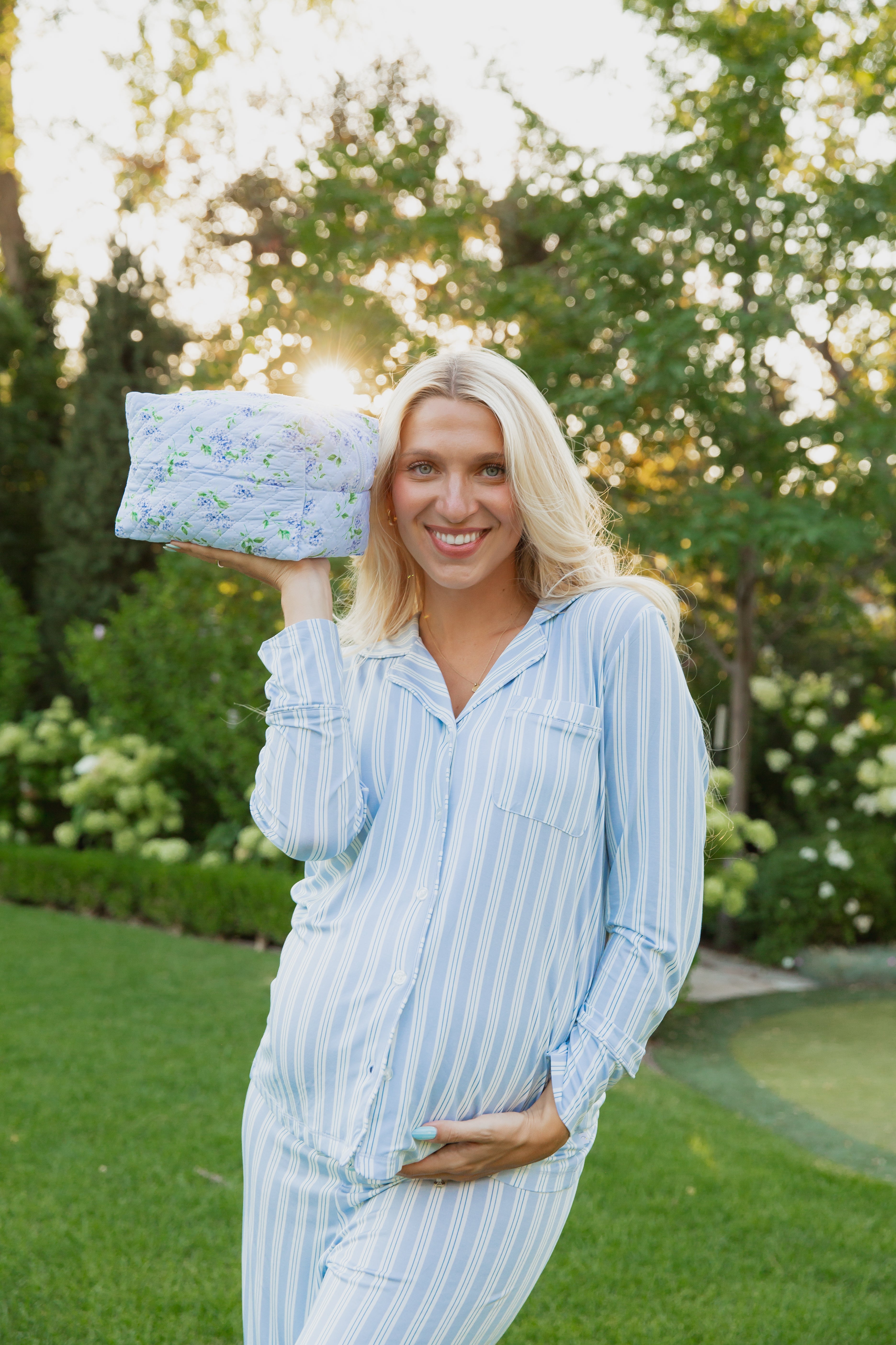 A smiling person in blue striped pajamas stands on the grass, holding a hydrangea / travel bag near their head with one hand resting on their belly. Sunlight and green trees fill the background, capturing a serene moment from the Goosebumps Collection.