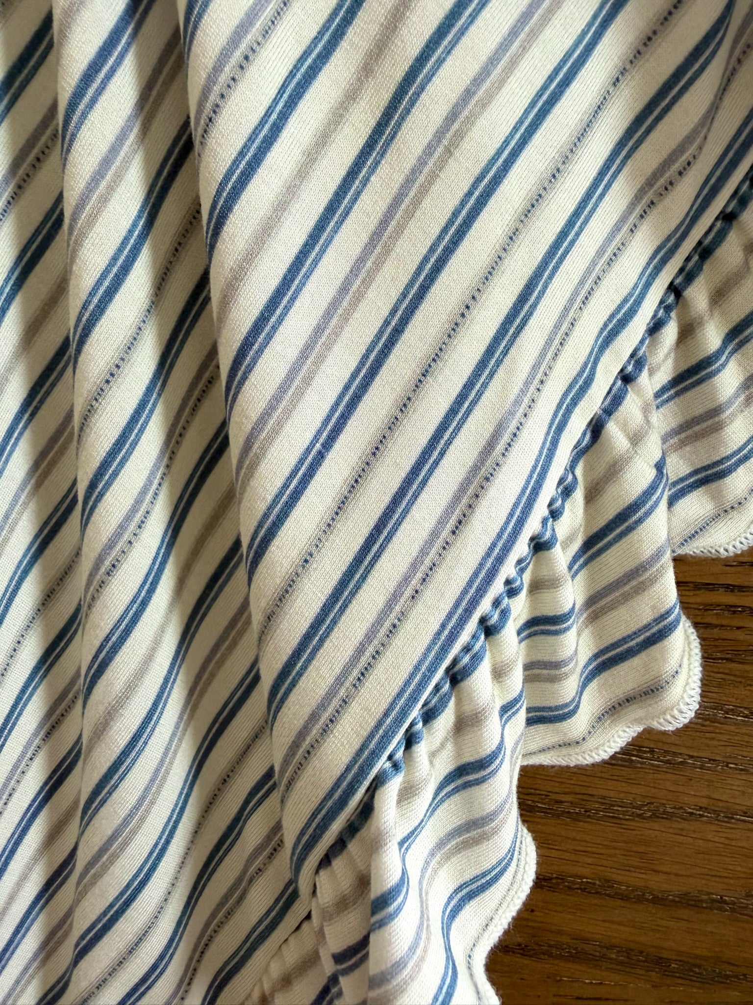 A close-up of the beach house / ruffle blanket features soft, scalloped edges and elegant diagonal blue and white stripes, draped over wood for a luxurious look.