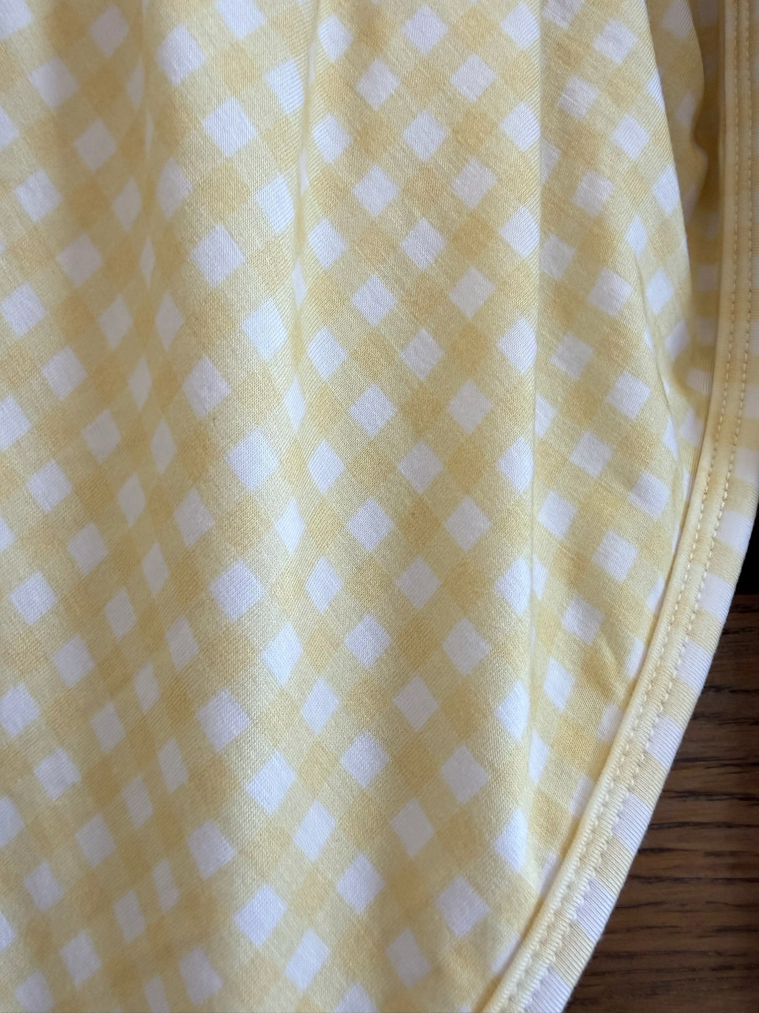 A close-up of the "sunshine" yellow gingham blanket with small checkered squares, showing a curved seam on the right edge and part of a wooden surface at the bottom right.