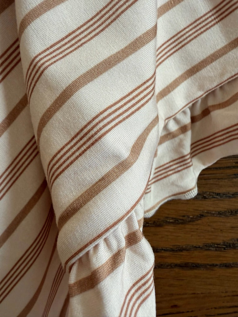 Close-up of the toast and honey / ruffle blanket—a luxurious, light fabric with brown diagonal stripes and a gathered ruffle edge—draped over a wooden surface with dark horizontal lines.