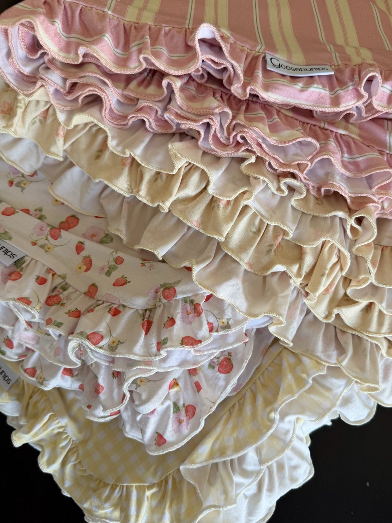 A close-up of five “freshly picked / ruffle” baby bloomers in pastel stripes, floral, strawberry, and gingham patterns, stacked on a strawberry print blanket.