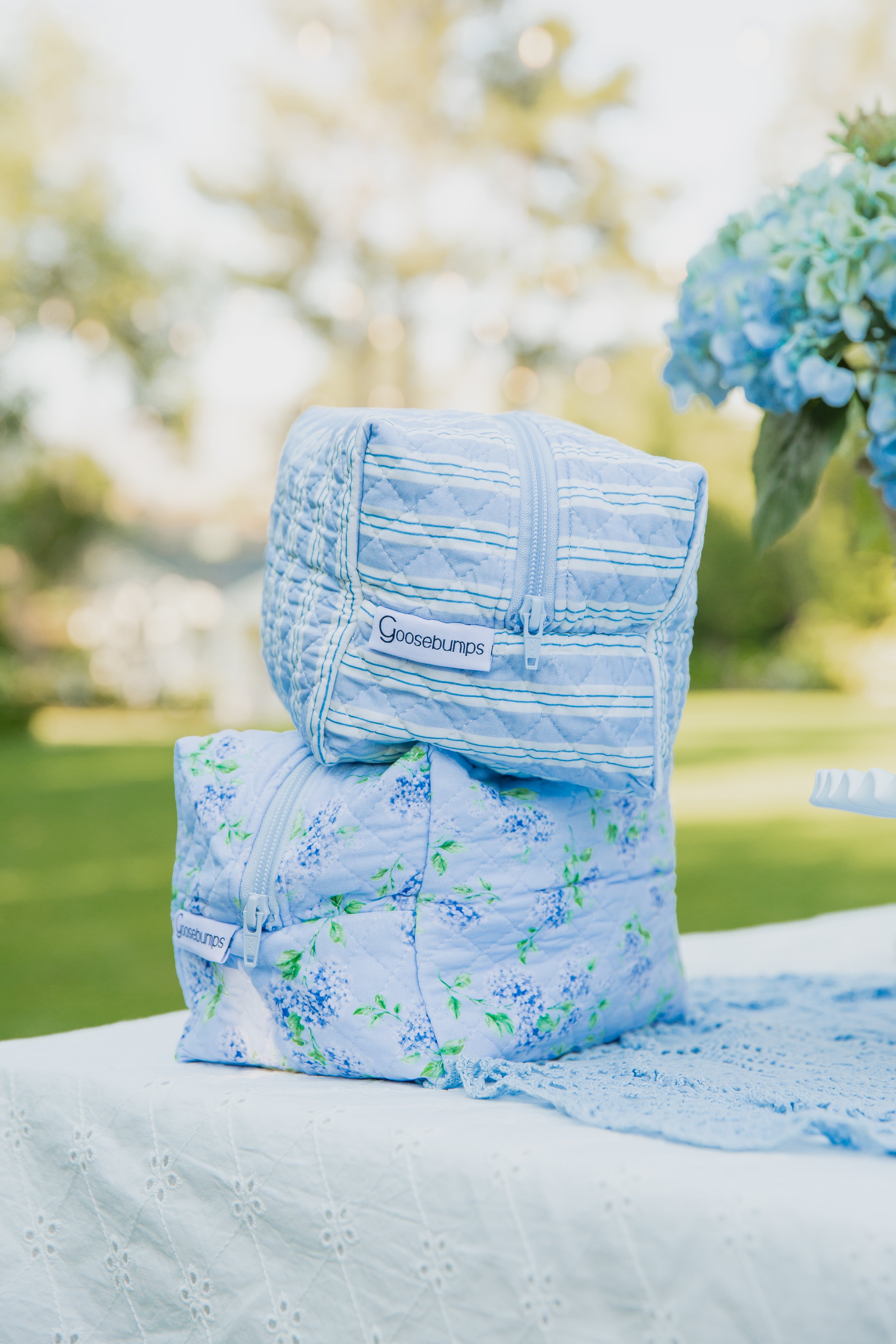 Two haven / travel bags in a pastel blue quilted design with Goosebumps tags are stacked on a white lace tablecloth outdoors, set against a backdrop of blurred greenery and blue flowers—perfect for weekend getaways or keeping your everyday essentials organized.