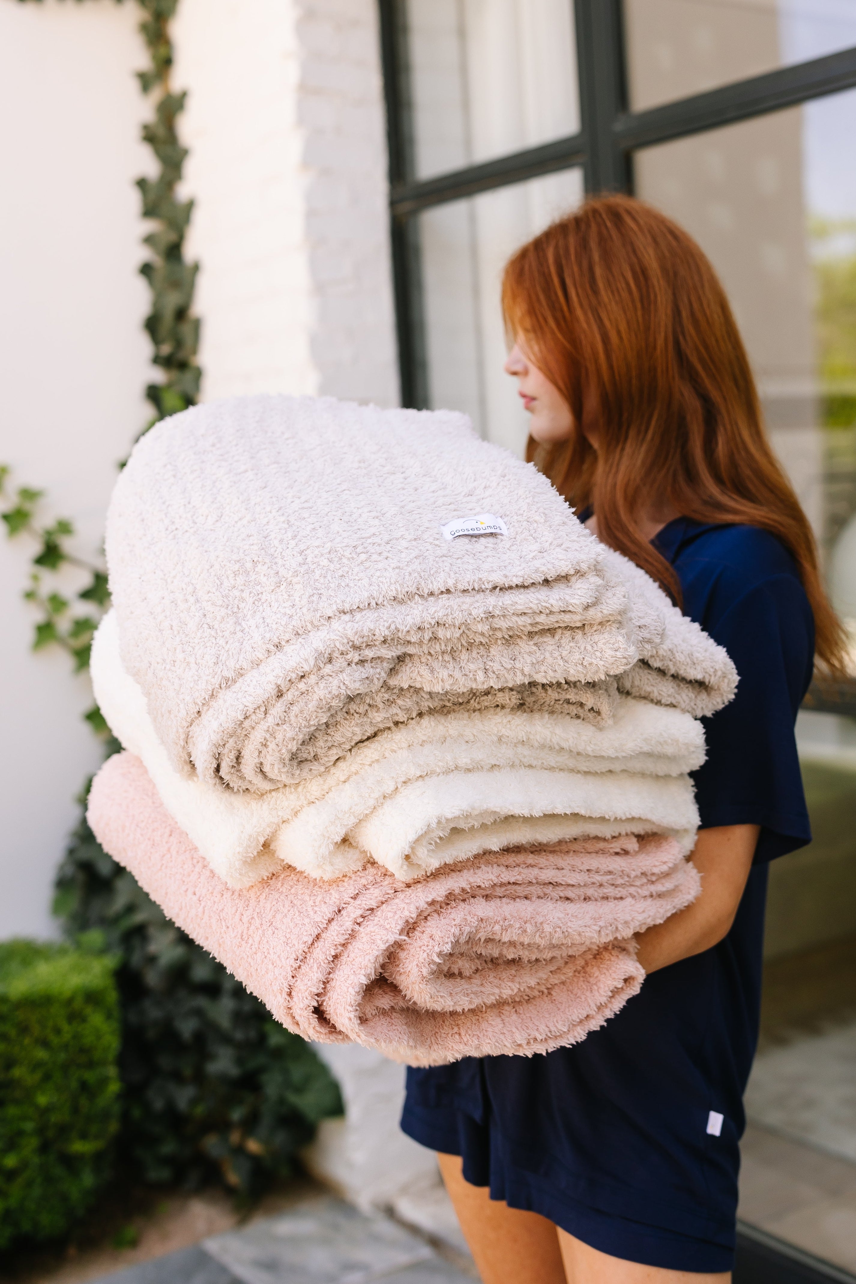 A person with long red hair in a navy blue outfit stands outside, holding a stack of ultra plush throws in beige, cream, and ballet slipper / plush. A brick wall with green ivy and glass doors are visible behind them.