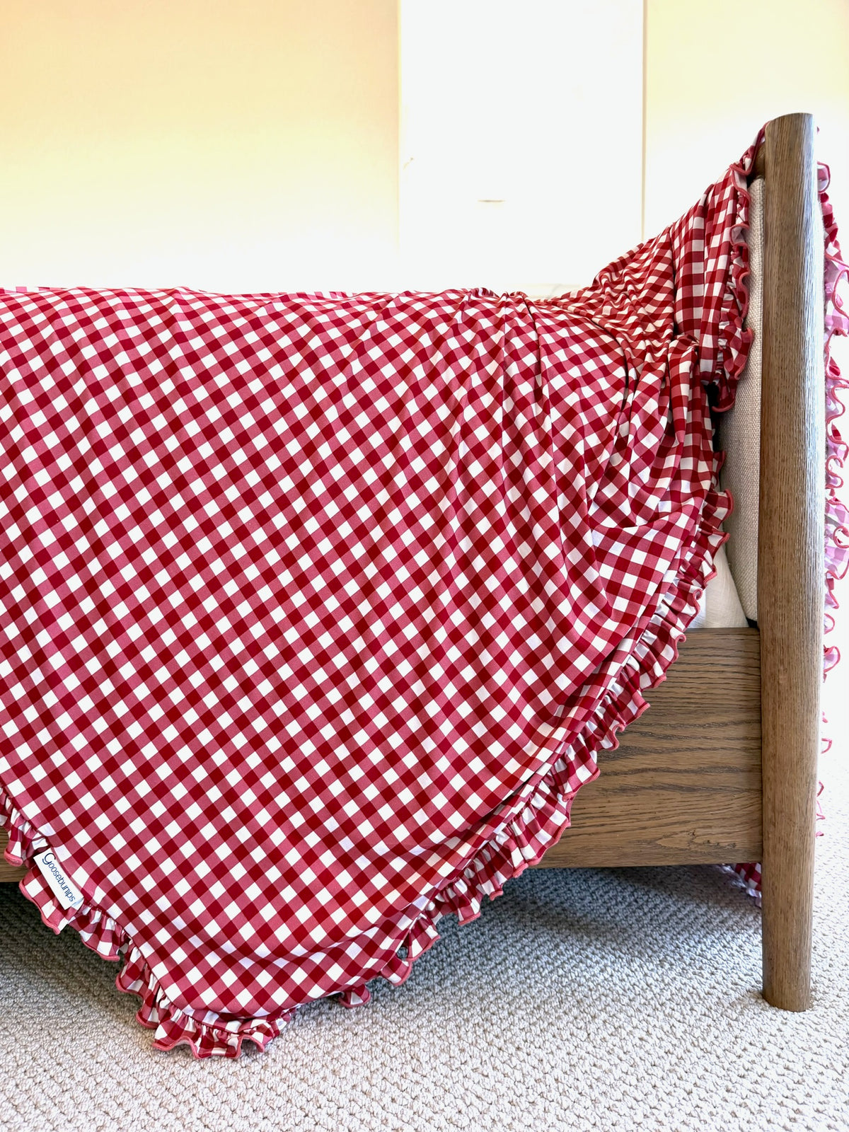 A modern, luxurious checkin’ it twice / ruffle blanket in red and white gingham with a ruffled edge draped over a wooden bed frame.