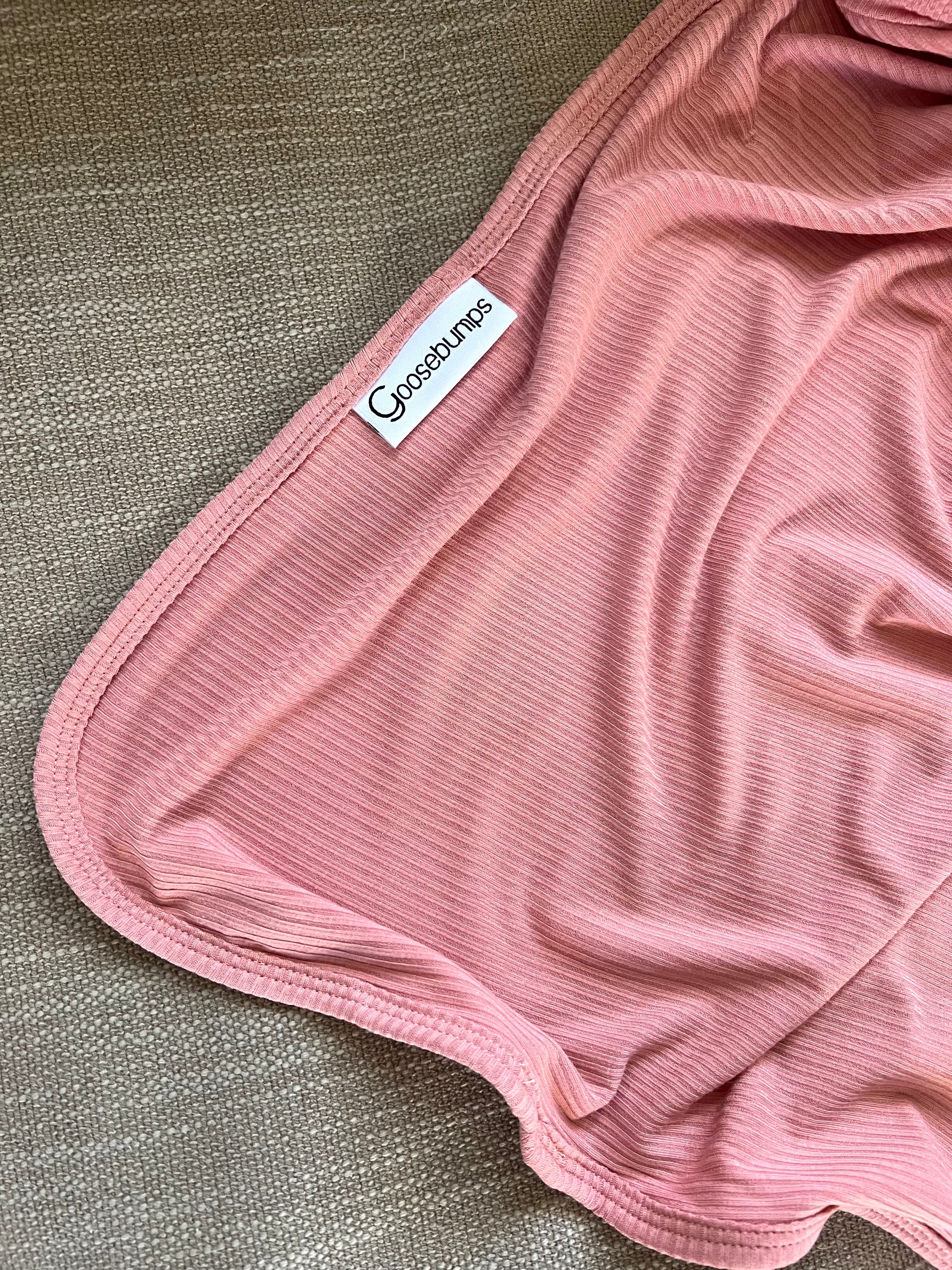 A close-up of the peony blanket, featuring soft ribbed pink fabric with fine stripes and a ConseWlynos label, luxuriously draped over a textured beige surface.