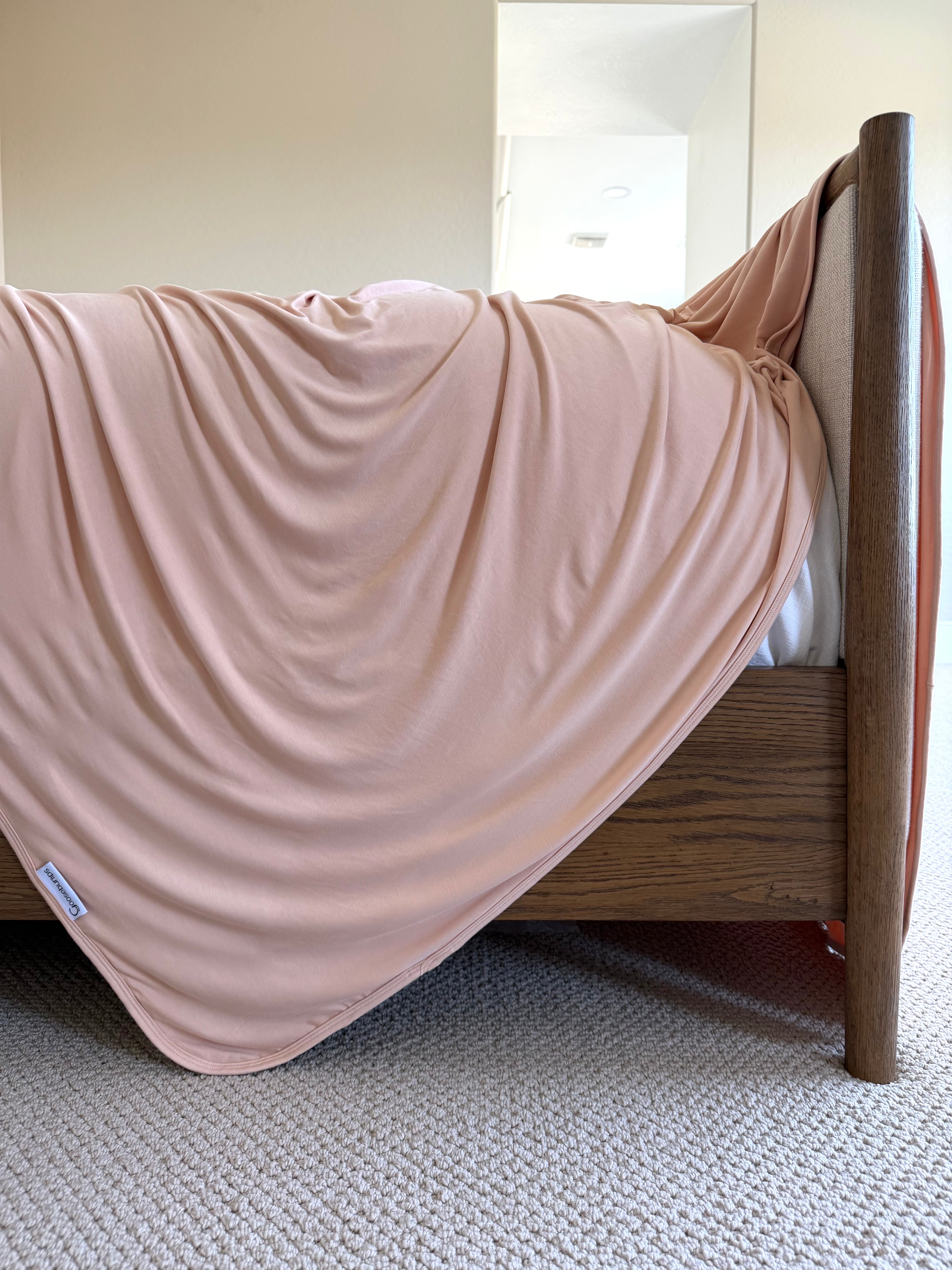A whisp blanket from the Lisa Wilson Goosebumps Collection in a peach hue drapes smoothly over the side of a wooden bed, offering the gentle embrace of an adult swaddle. The soft bamboo fibers blend perfectly with the beige carpet and softly lit, neutral-toned room in the background.