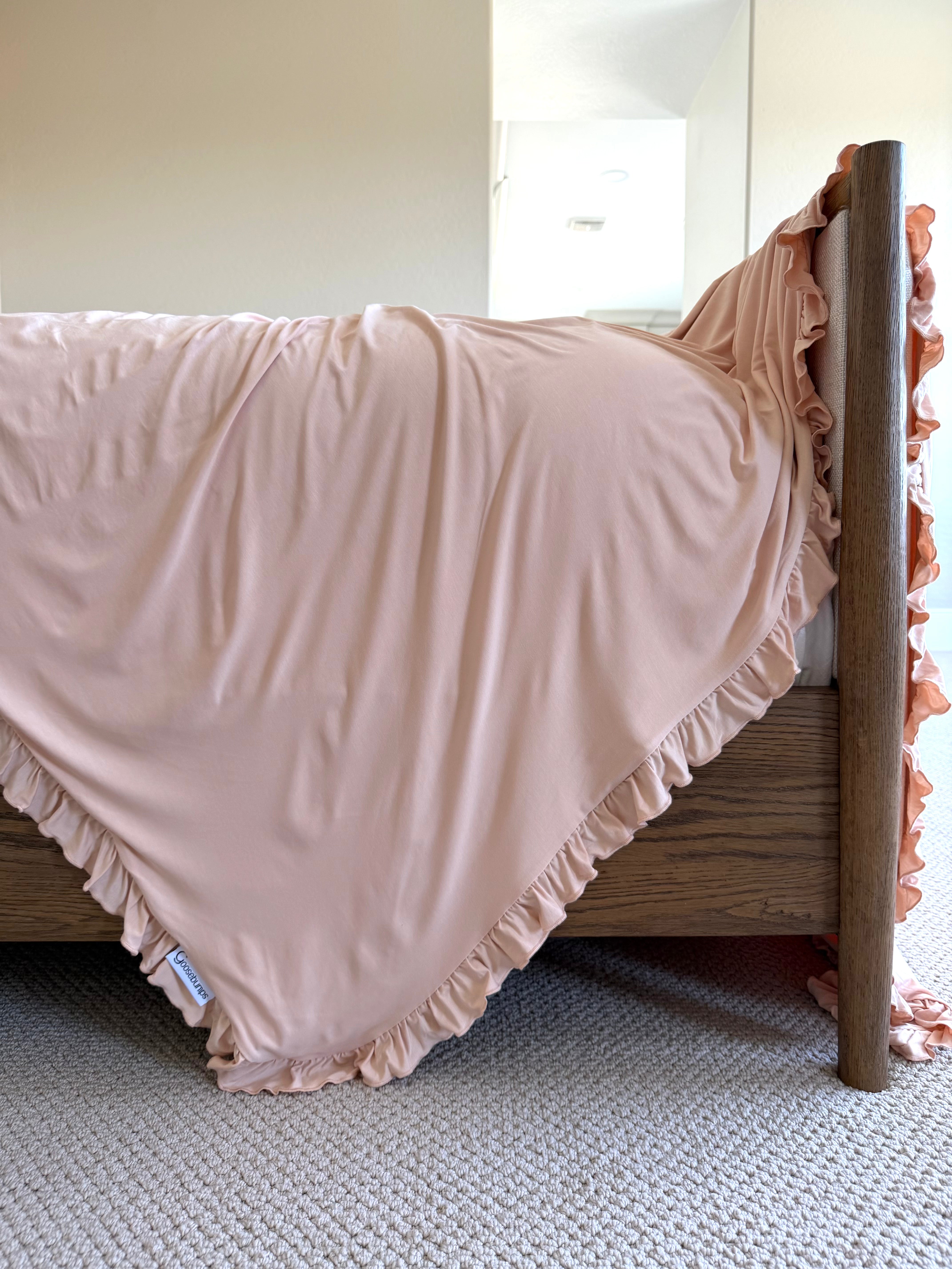 A wooden bed is draped with the whisp / ruffle—a pale pink blanket adorned with elegant ruffle trim—which cascades over the side and grazes a light-colored carpeted floor in a softly lit bedroom.