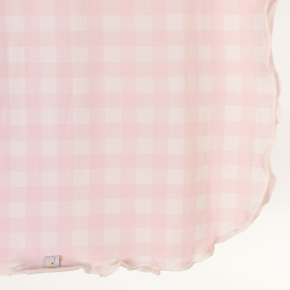The xo / swaddle is a pink and white gingham blanket with scalloped edges, draped softly. Made from gentle viscose fabric, it features a small tag at the bottom left corner.