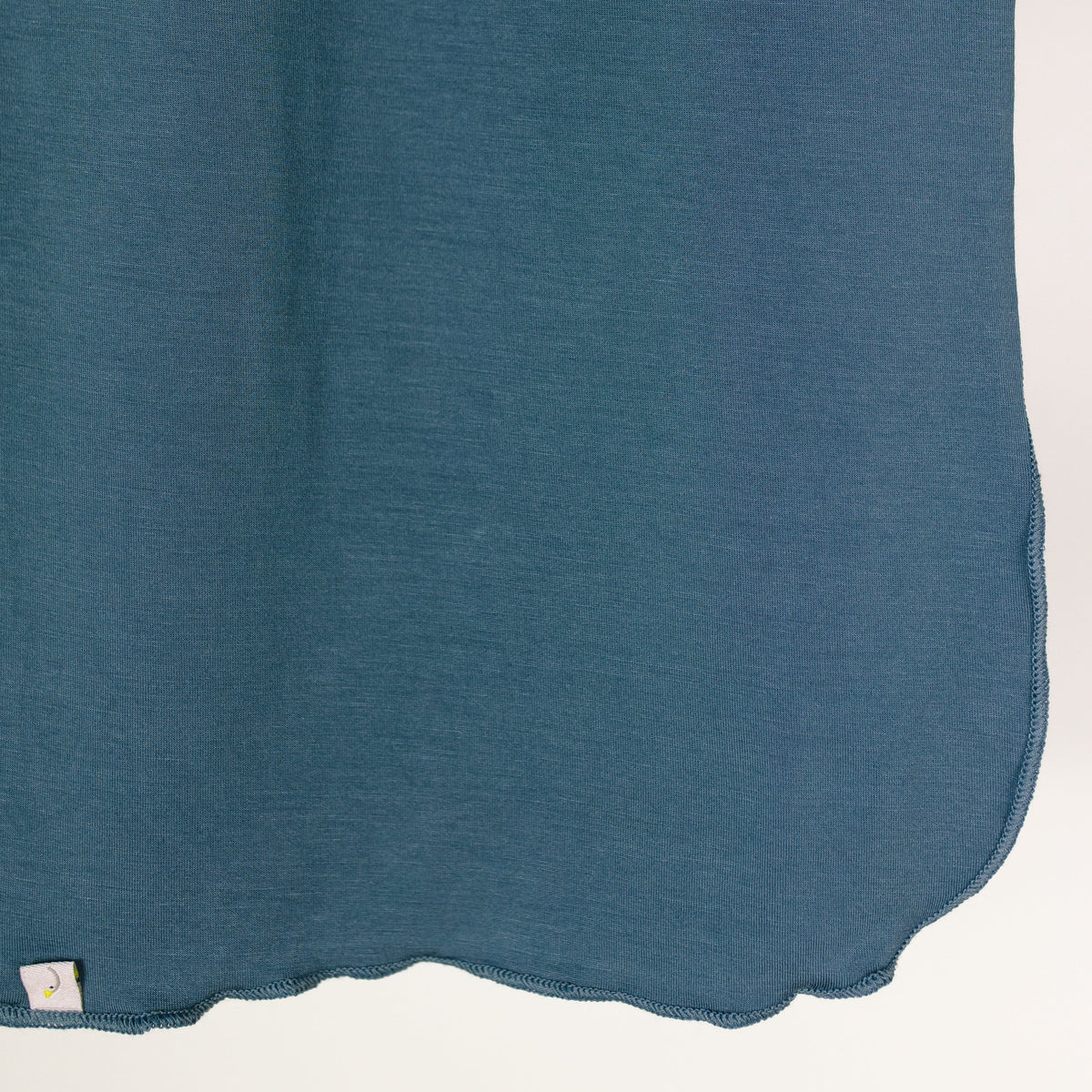 A close-up of the tidal wave / swaddle showcases its teal fabric, delicate scalloped hem, and small stitched tag near the edge, set against a plain white background and highlighting the exquisite craftsmanship of this single-layered swaddle.