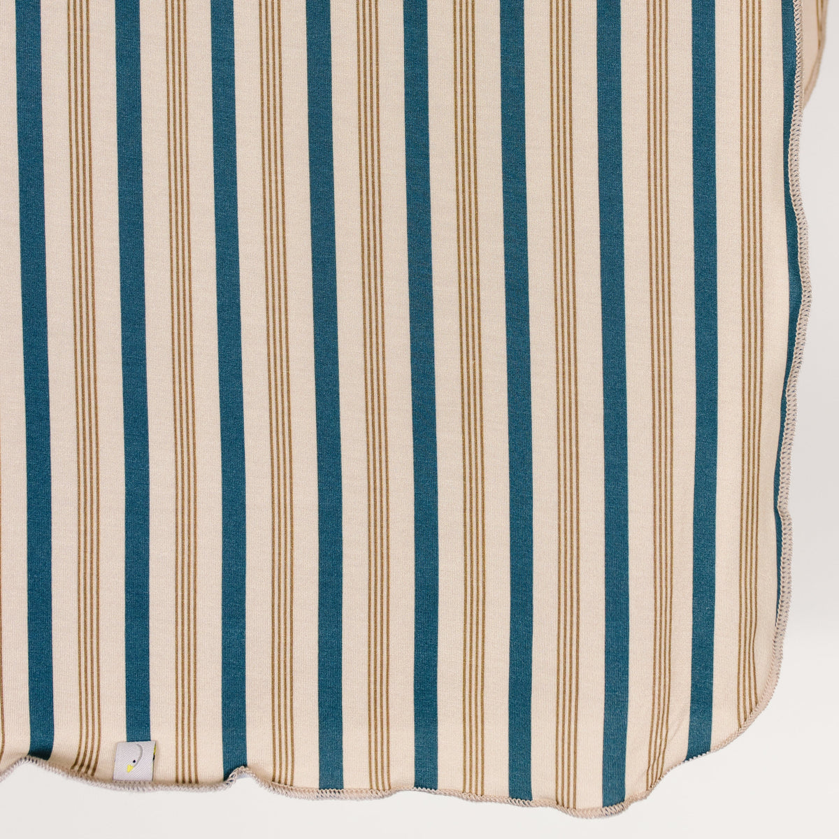 The “shoreline / swaddle” features vertical tan and indigo stripes, a scalloped edge, and a small tag near the bottom. Made from single-layered swaddle material, it is displayed against a plain white background.