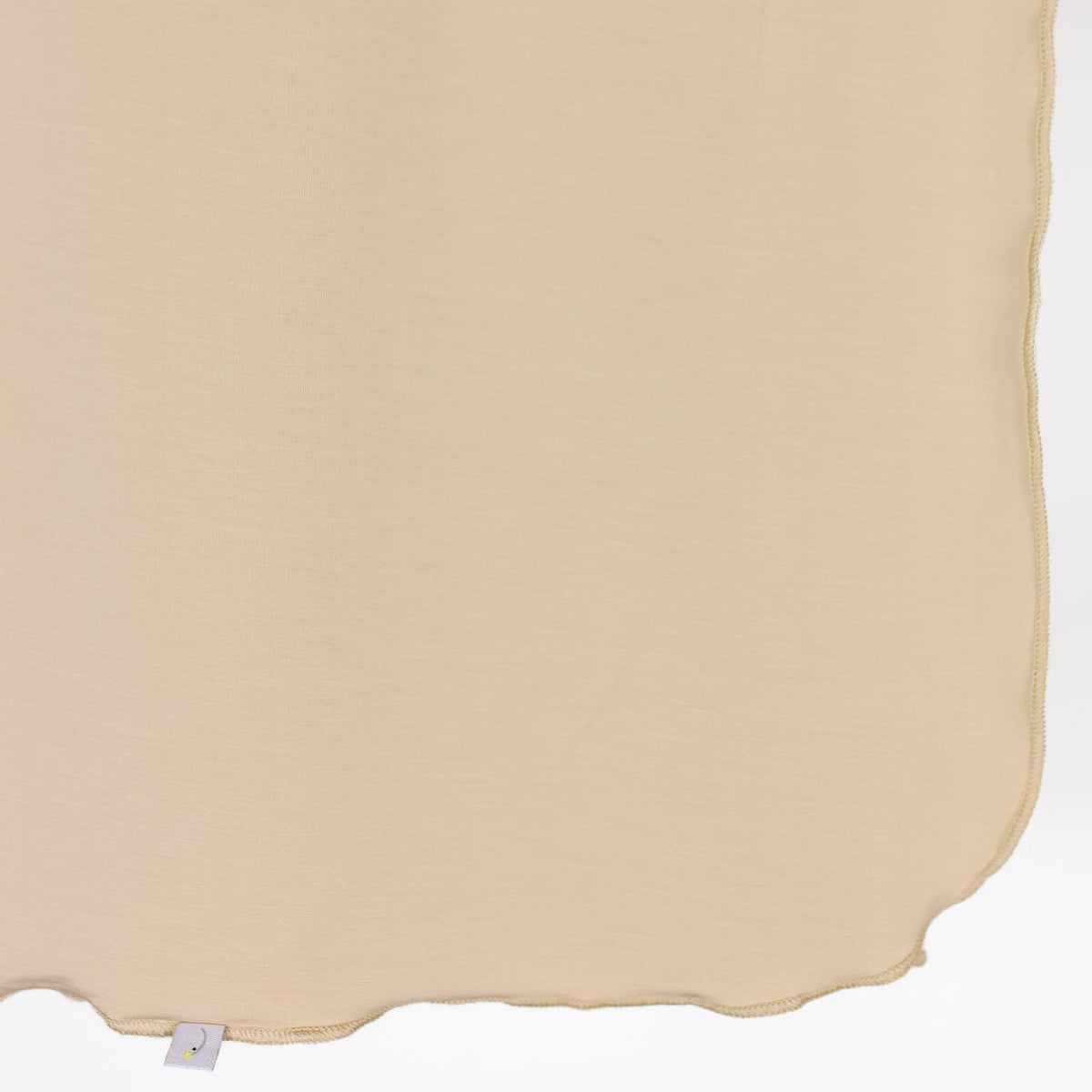 A vanilla cream swaddle hangs against a white background, featuring a wavy hem and a small white tag with a purple detail attached near the bottom edge.